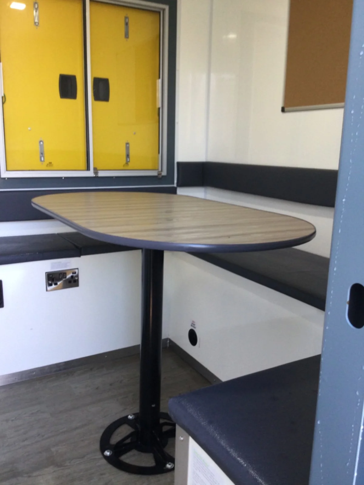 Empty seating area with a wooden table attached to a black metal pedestal, dark bench with black cushioned backrest, and yellow lockers in the background.