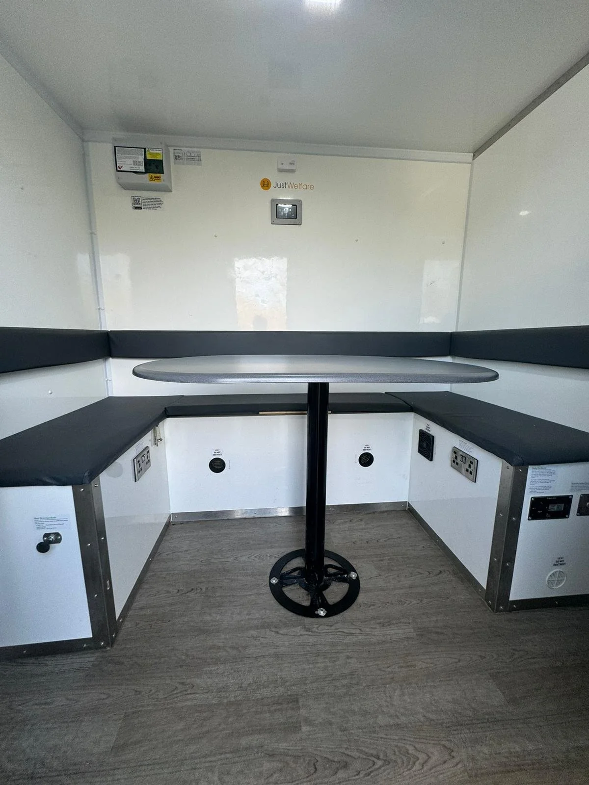 Compact white room with black cushioned bench seat along three walls, a round table in the center, and electrical outlets and controls on the walls and floor.