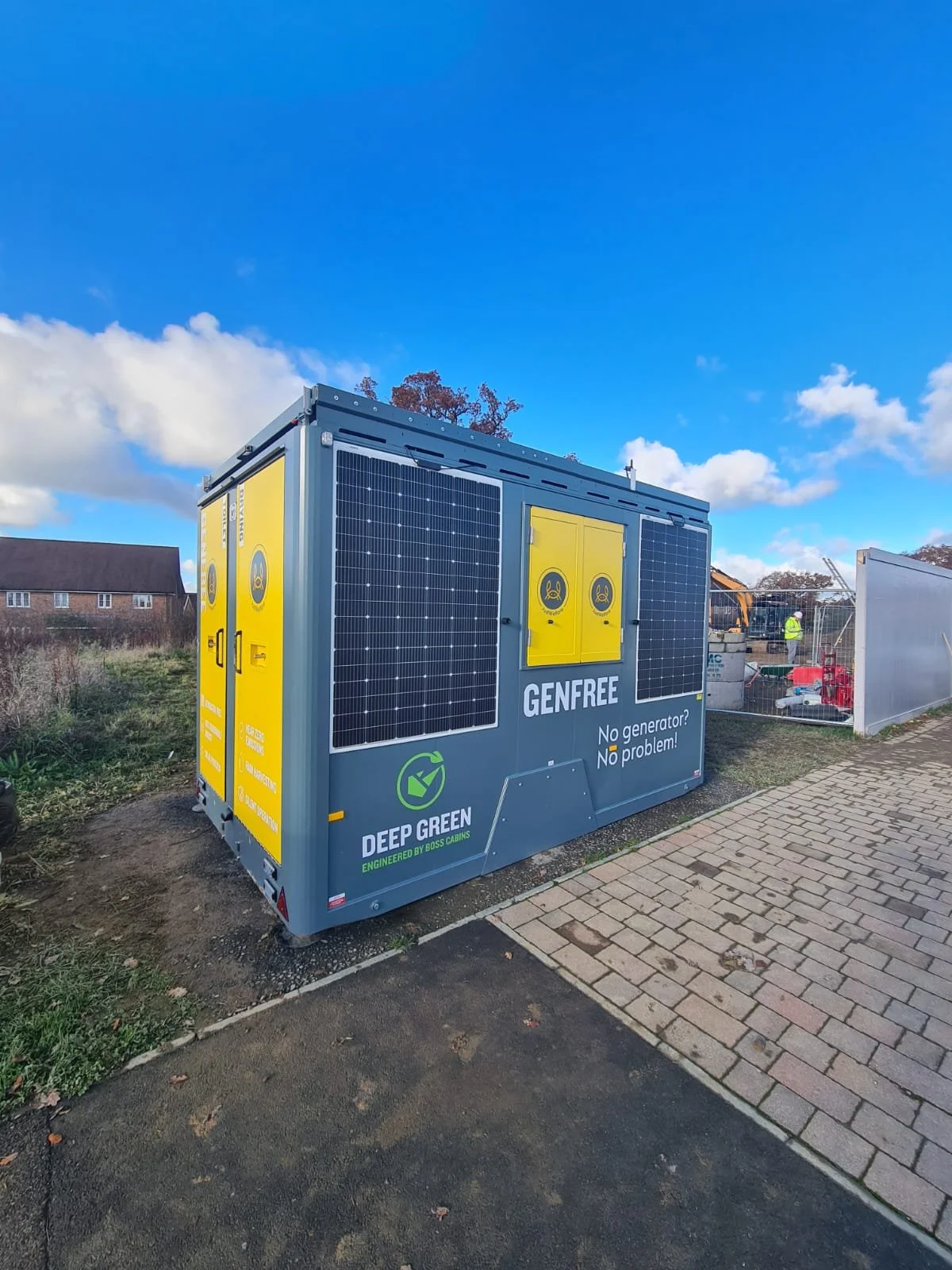 A solar-powered generator with yellow doors, black solar panels, and branding that says 'GENFREE' and 'DEEP GREEN,' situated outdoors near a sidewalk and construction site under a blue sky with clouds.