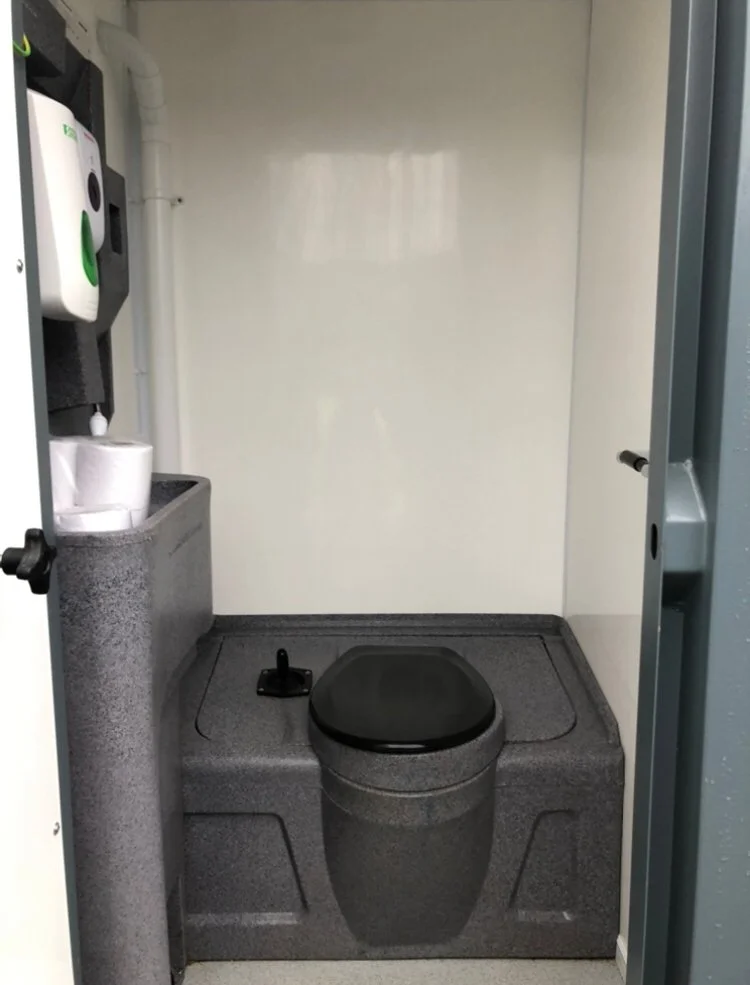 Small, gray, portable toilet with a black seat and lid, a black toilet brush on the side, and a small sink area with a paper towel dispenser and rolls of toilet paper, set in a compact, enclosed space.