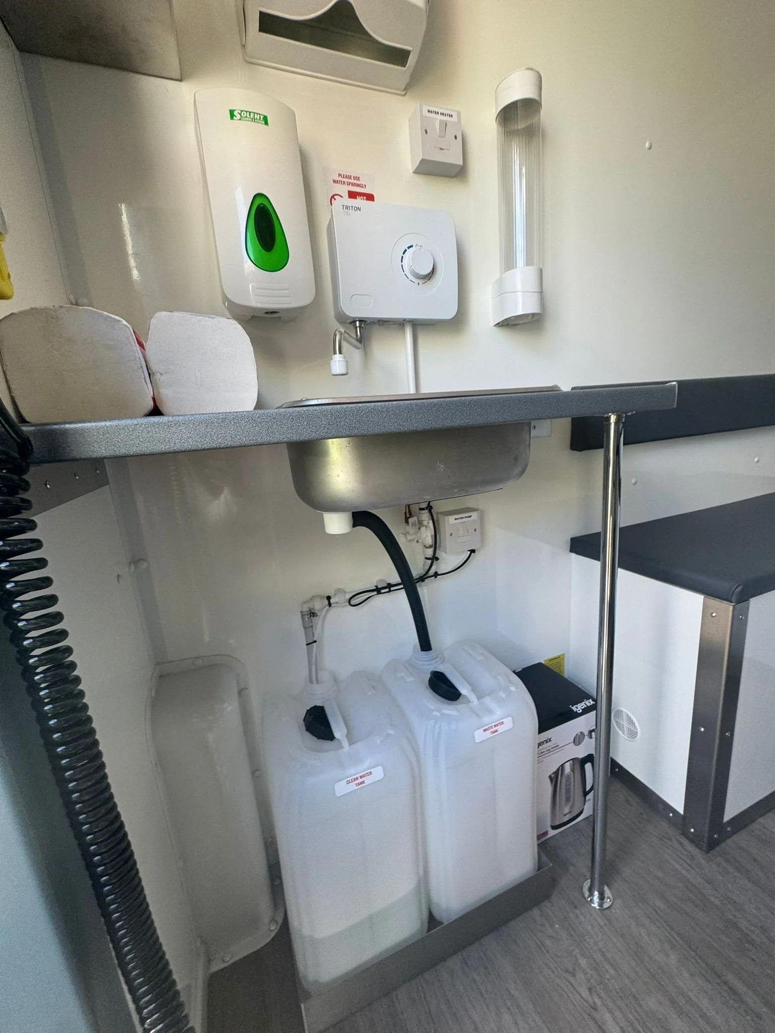 A hospital or medical room sink with two white water containers underneath, disinfectant and soap dispensers on the wall, and a water heater.