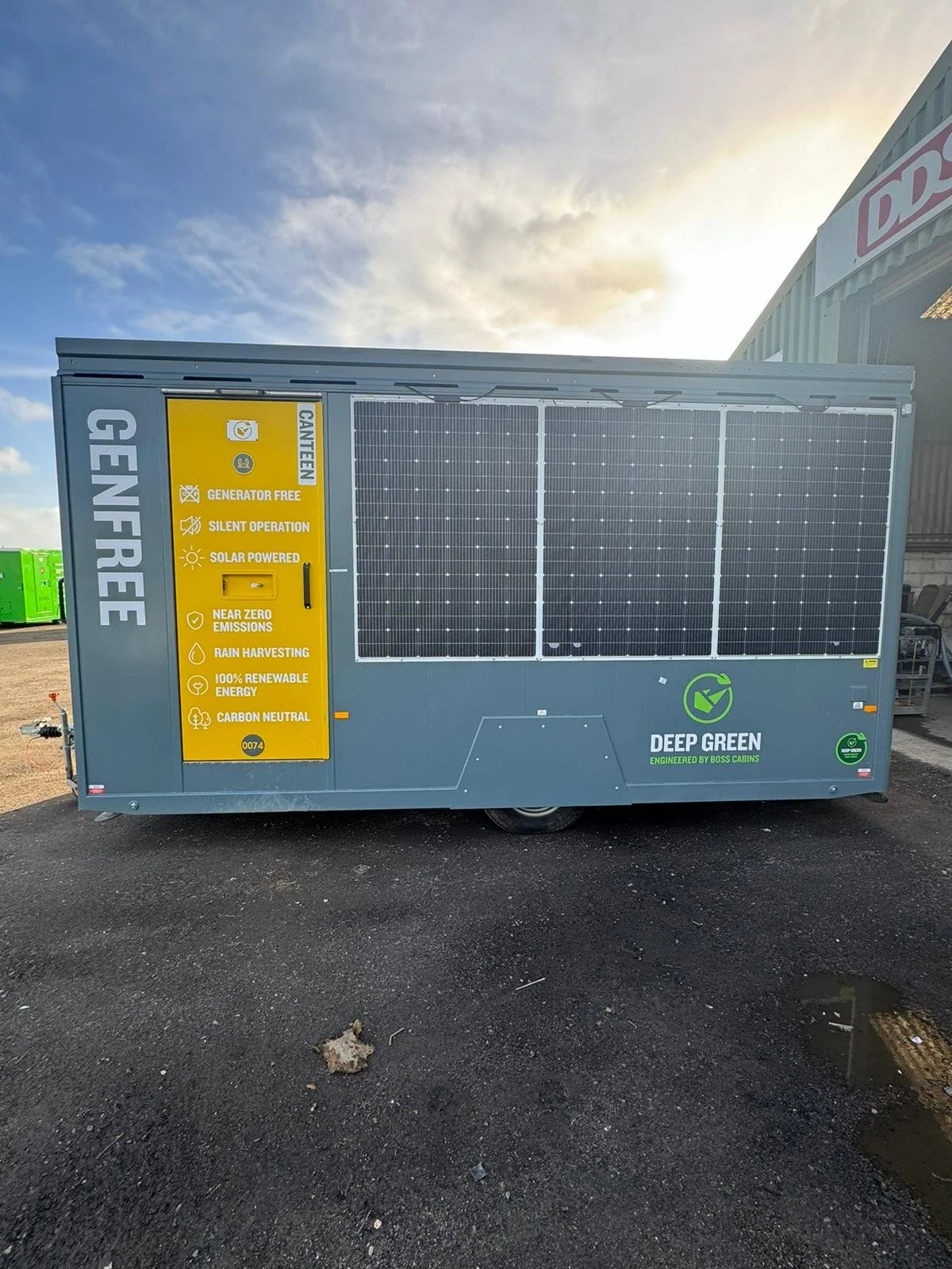 A solar-powered generator trailer with yellow and green signage, situated outdoors on asphalt near a building in daytime with partly cloudy sky.