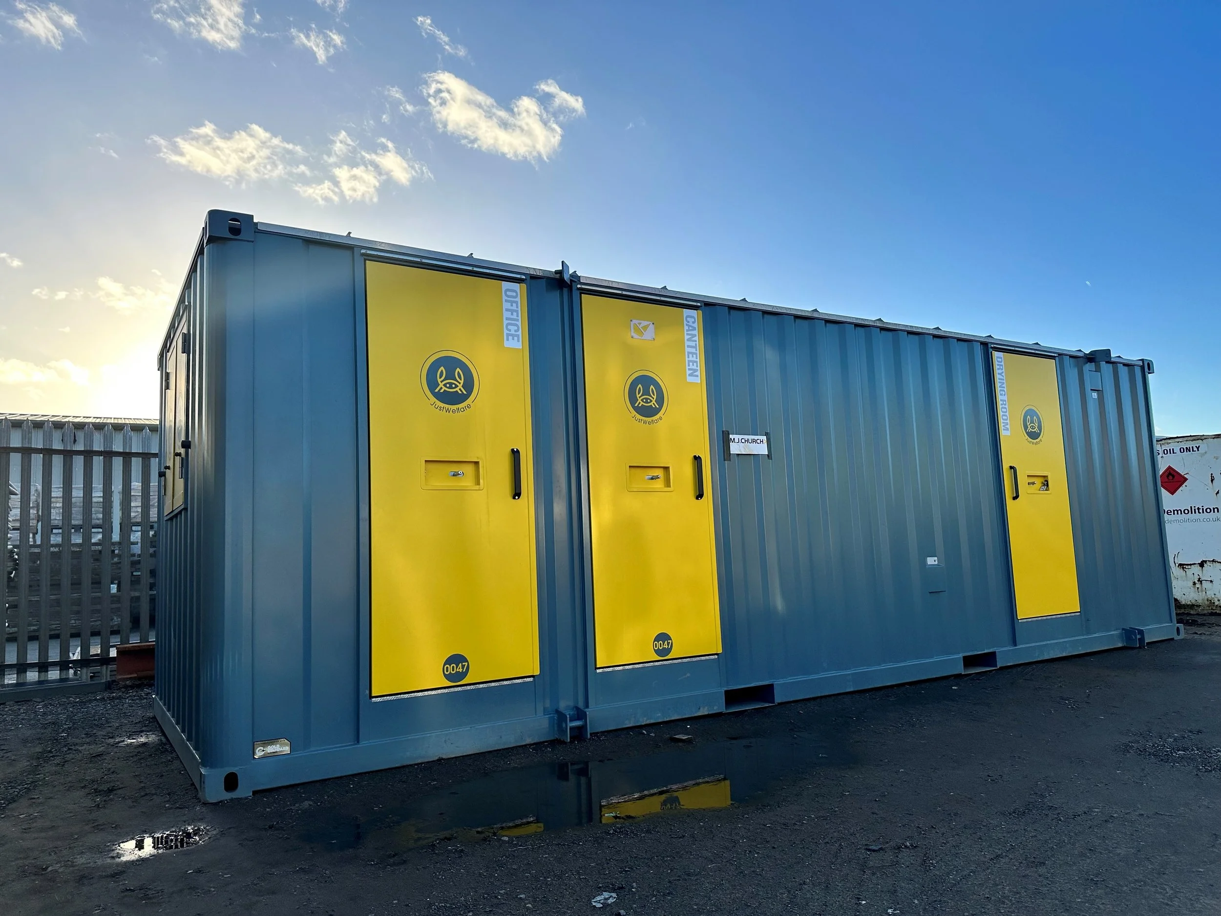 A grey shipping container with bright yellow doors labeled for various utilities, situated outdoors on a paved ground, with a fence and a blue sky with clouds in the background.