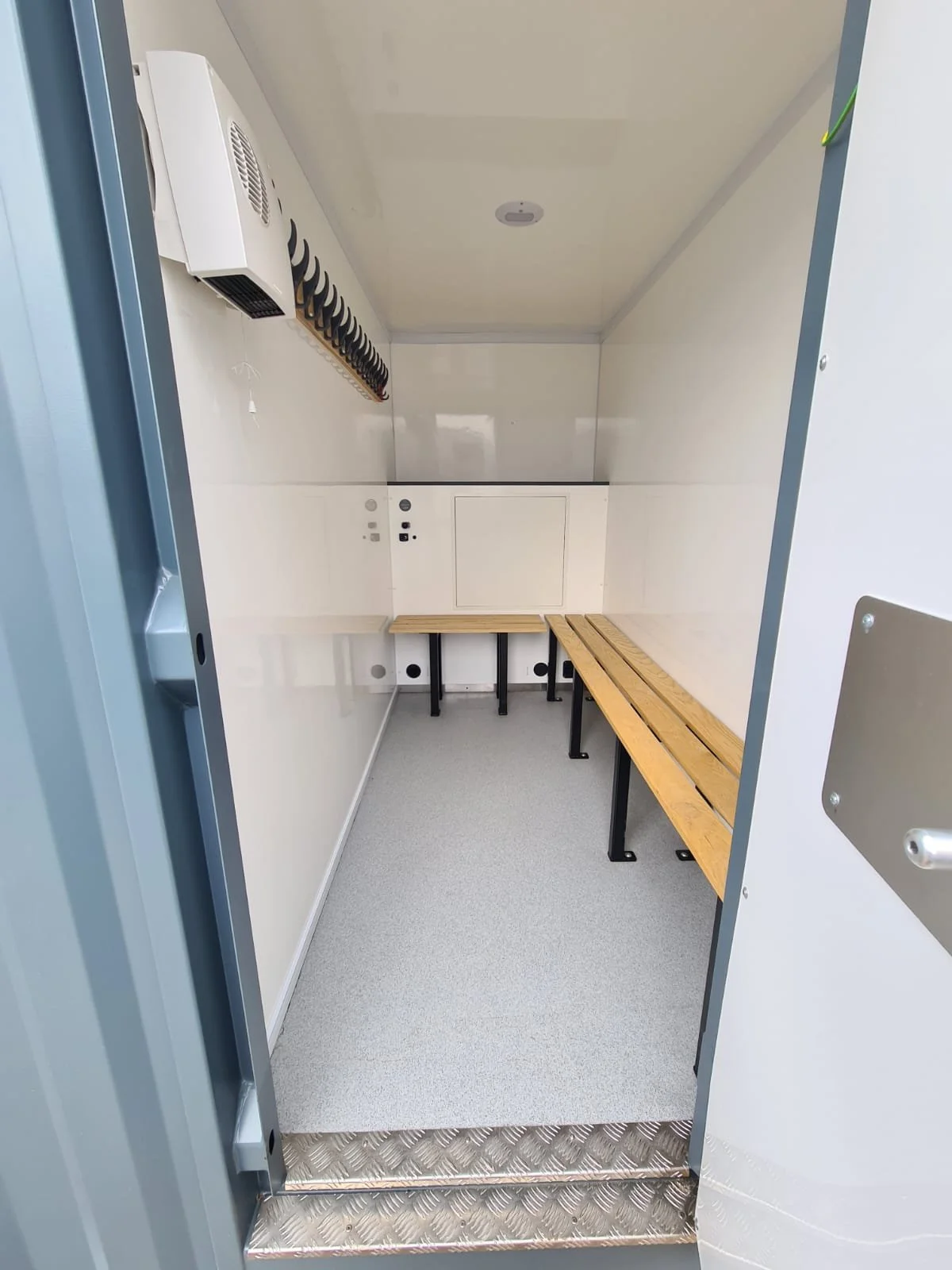 Inside a small, clean, empty waiting area with wooden benches, a white wall with electrical outlets, a wall-mounted heater, and a ceiling light fixture.
