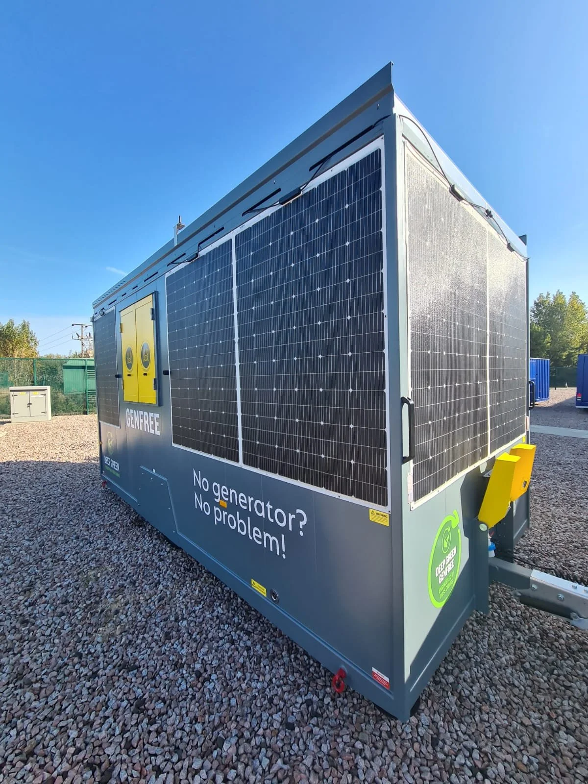 A large portable solar generator with solar panels on the sides, placed on gravel outside under a clear blue sky. The generator has branding and text that says 'No generator? No problem!' and features a green sticker for Deep Green.