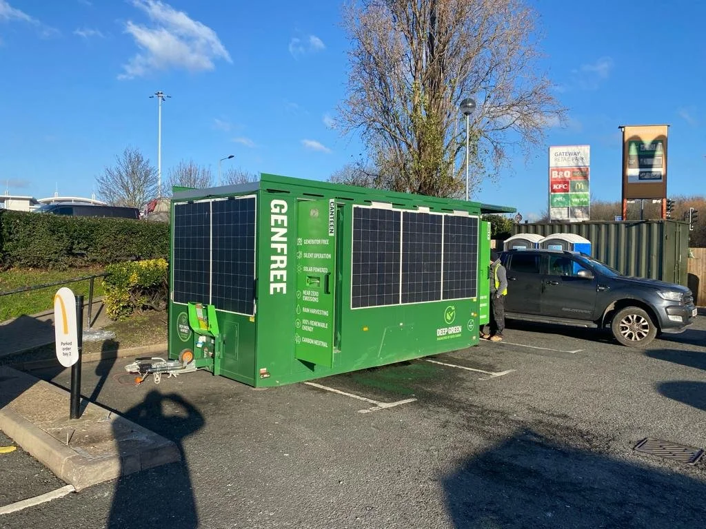Green solar-powered container in a parking lot labeled Deep Green, with solar panels on its sides, surrounded by parked cars, trees, and signs for other businesses.