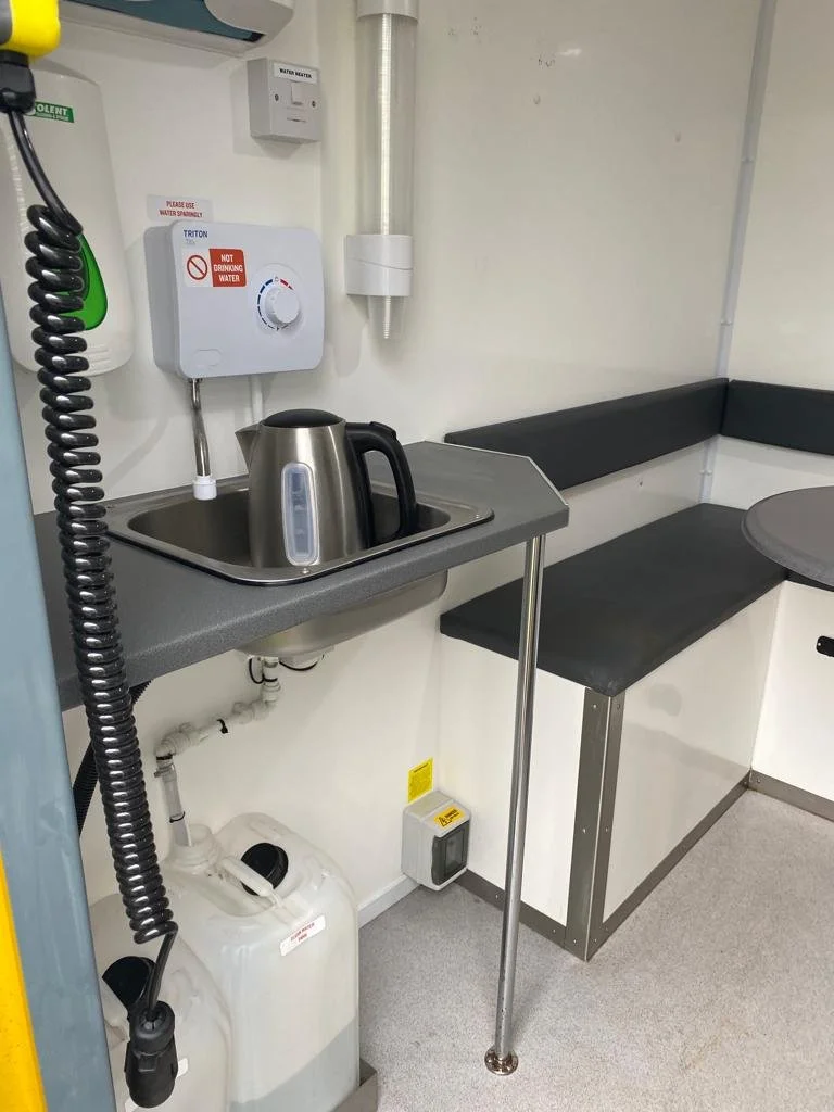 A small corner of a medical or laboratory room with a sink, electric kettle, water filtration system, and black bench seating.