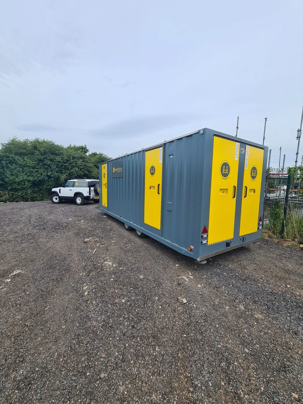 A mobile police station container with yellow doors and the police logo, parked on a gravel lot with a white vehicle in the background and some trees on the horizon.