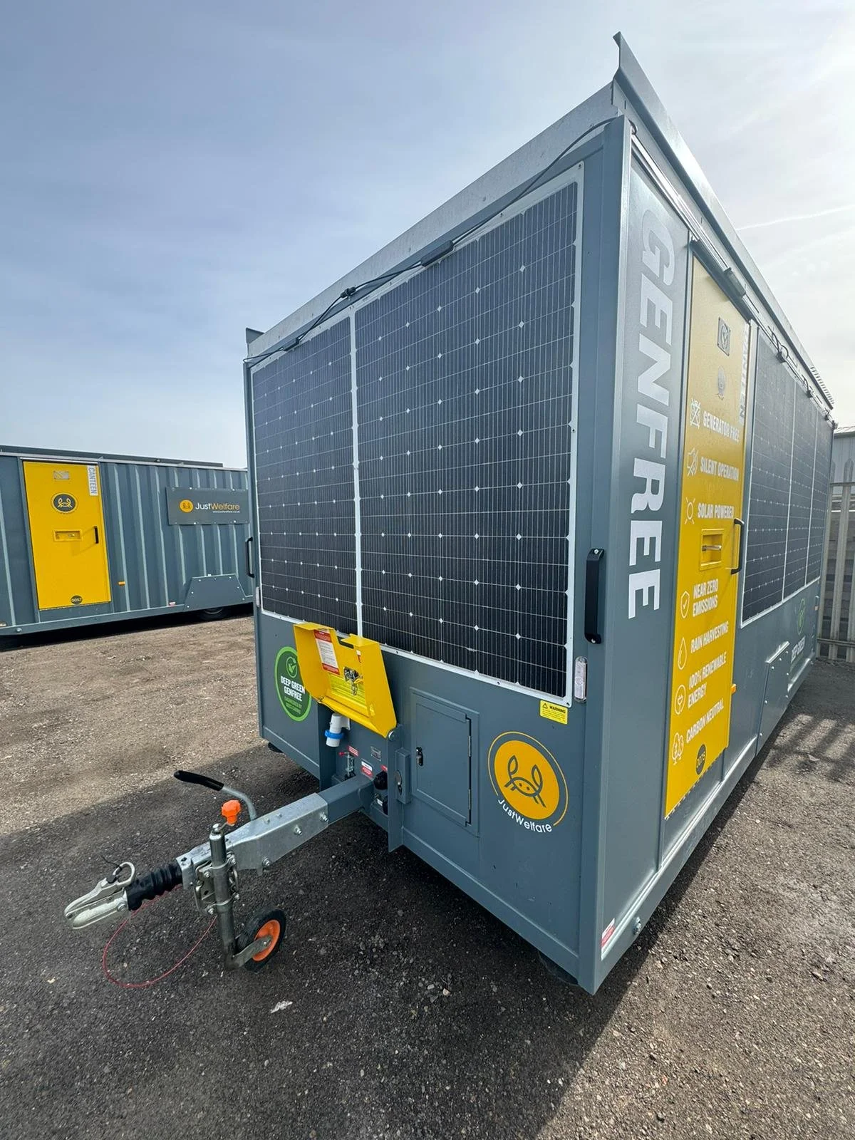 A large portable power generator with solar panels on the side, labeled 'GENFRE,' positioned outdoors on a gravel surface, with another MWT generator in the background, under a clear sky.
