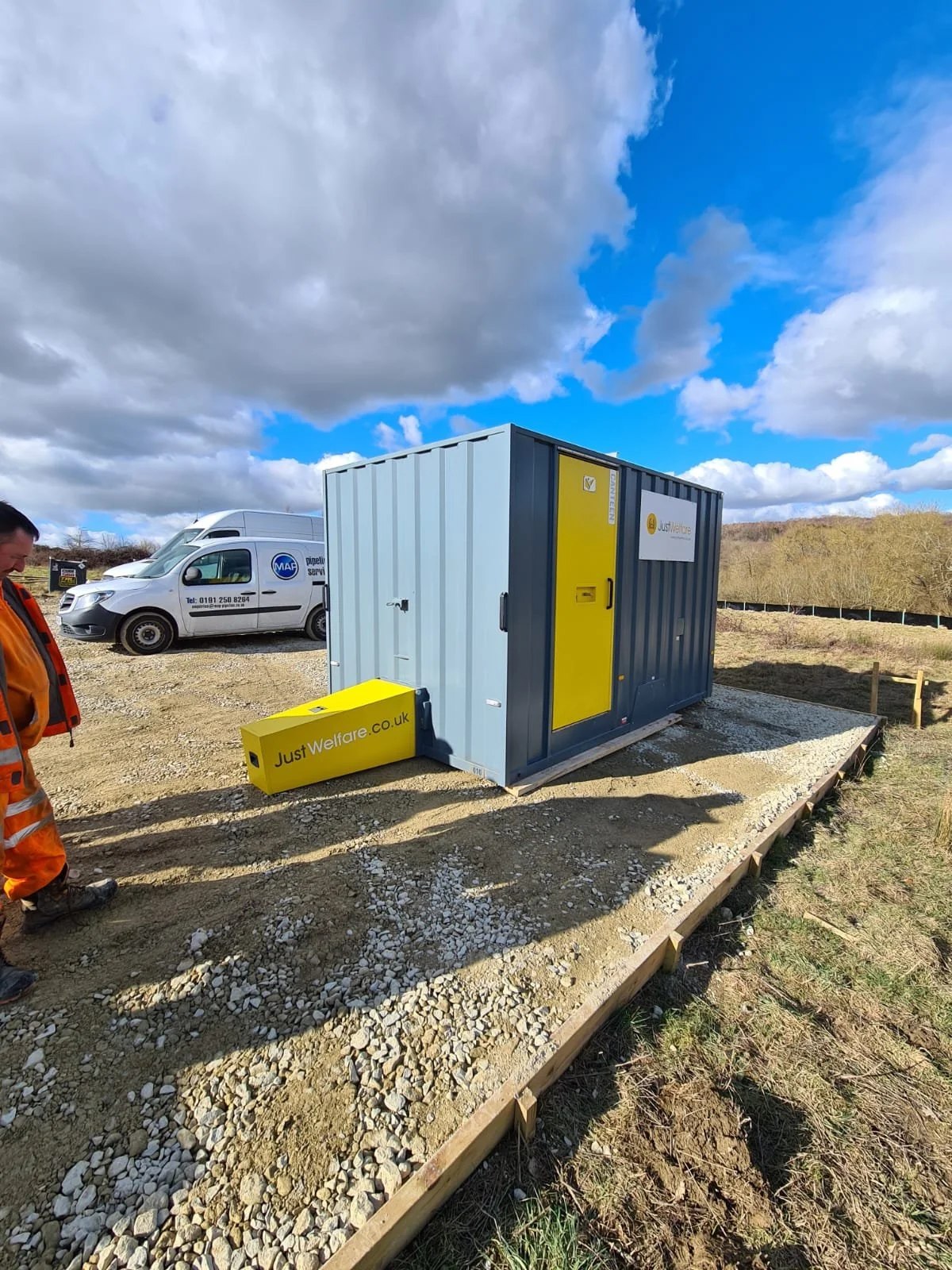 A portable welfare unit with a gray structure and yellow doors, seated on a gravel foundation outdoors under a partly cloudy sky. A man in an orange high-visibility jacket and pants stands nearby, with vehicles in the background including a white van