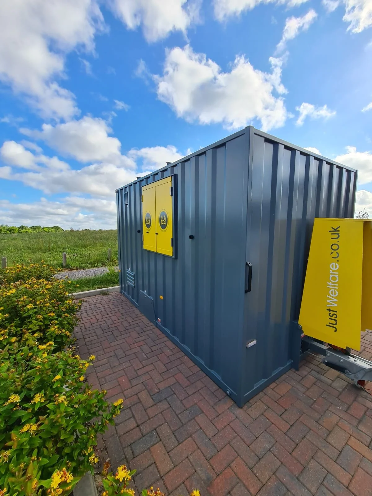 A large blue generator with yellow warning signs on the door, located on a brick path beside some yellow flowering bushes under a partly cloudy sky.