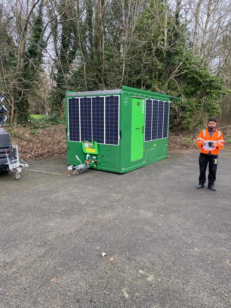 Green mobile solar-powered generator with solar panels, man in orange safety vest holding a tablet, outdoors on paved surface near trees.