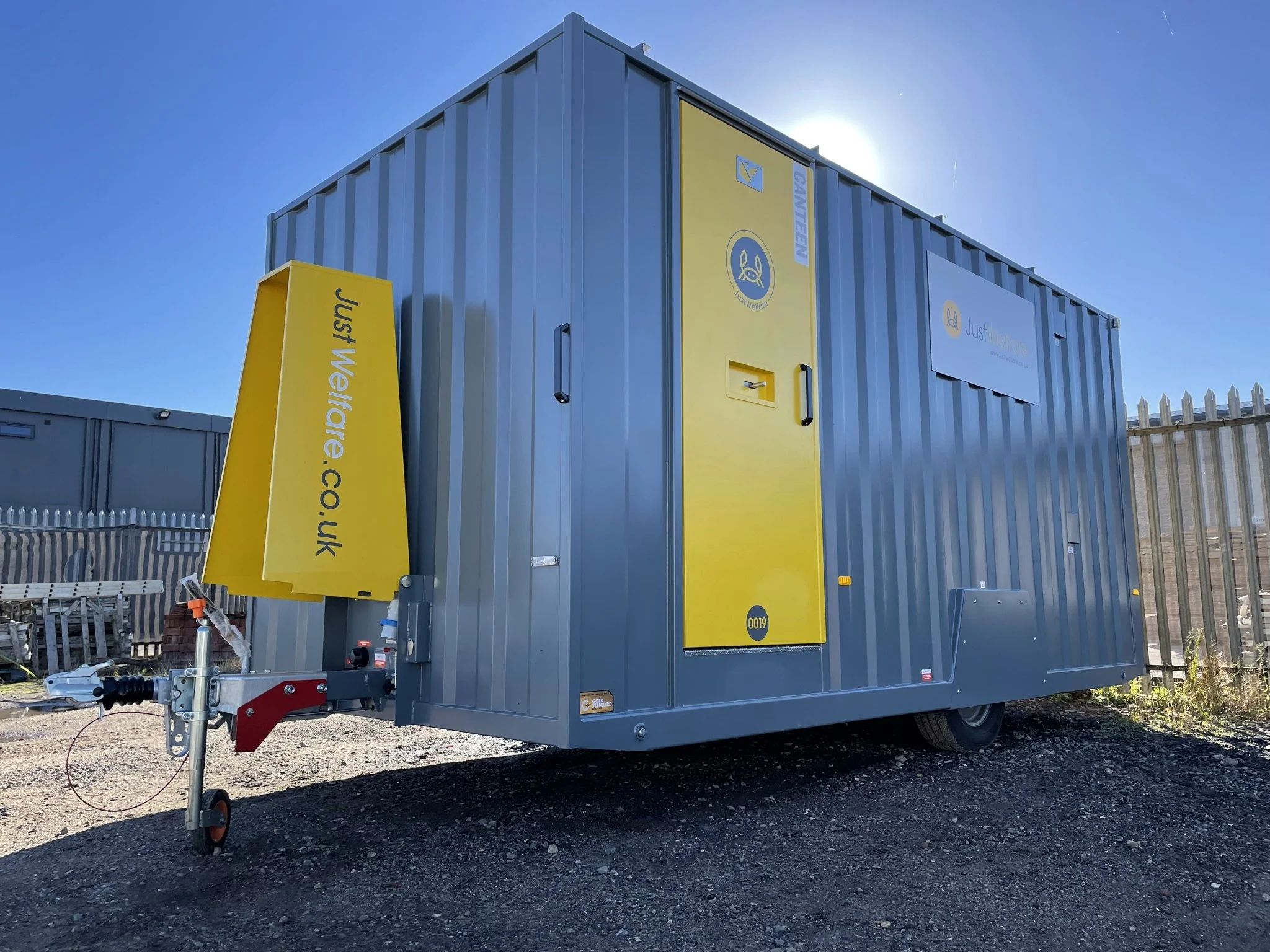 A large grey portable toilet trailer with yellow doors and signage, parked on gravel, with a fenced outdoor area in the background and the sun shining behind it.