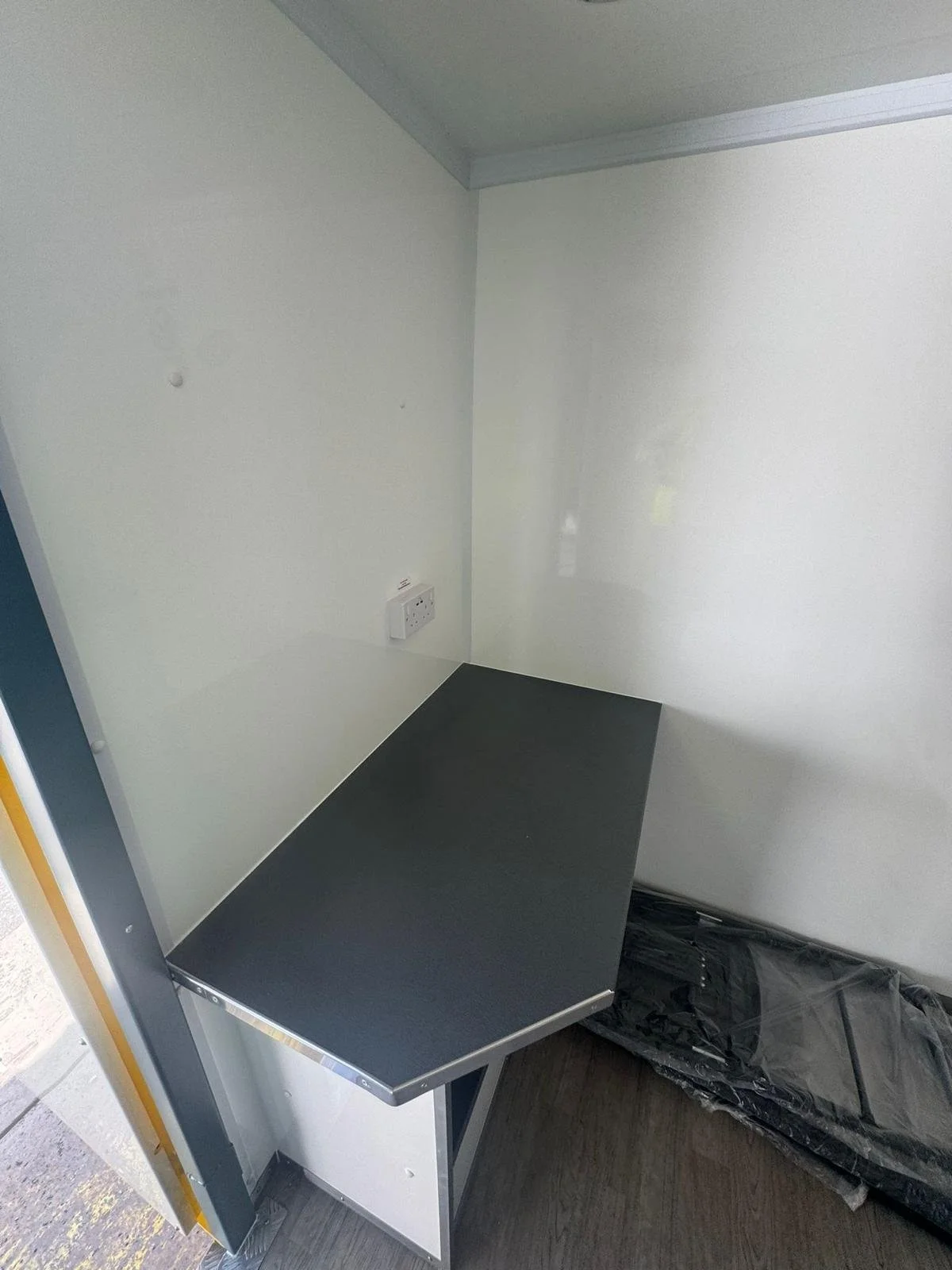 Empty small room or booth with blank white walls, a black countertop or desk, and a power outlet on the wall.