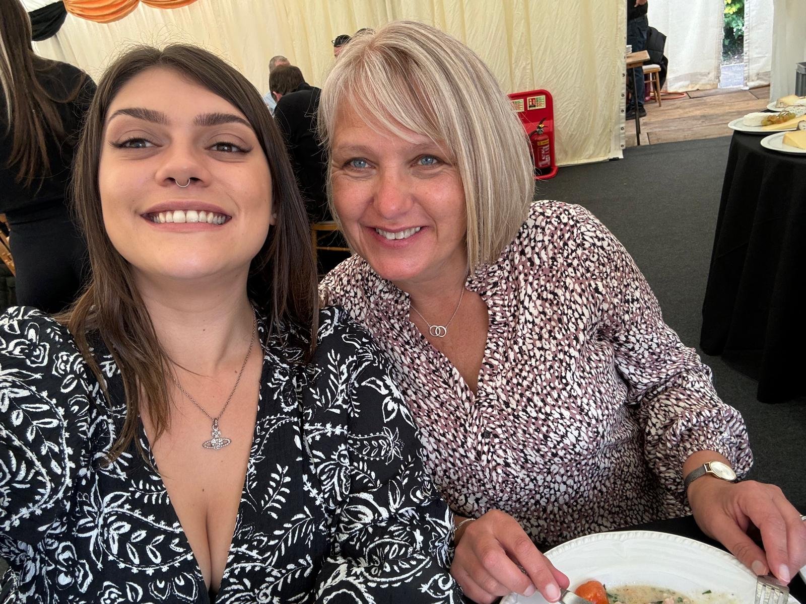 Two women smiling and taking a selfie at a gathering. They are seated at a table with plates of food and the background shows other people, tables, and a partially open door.