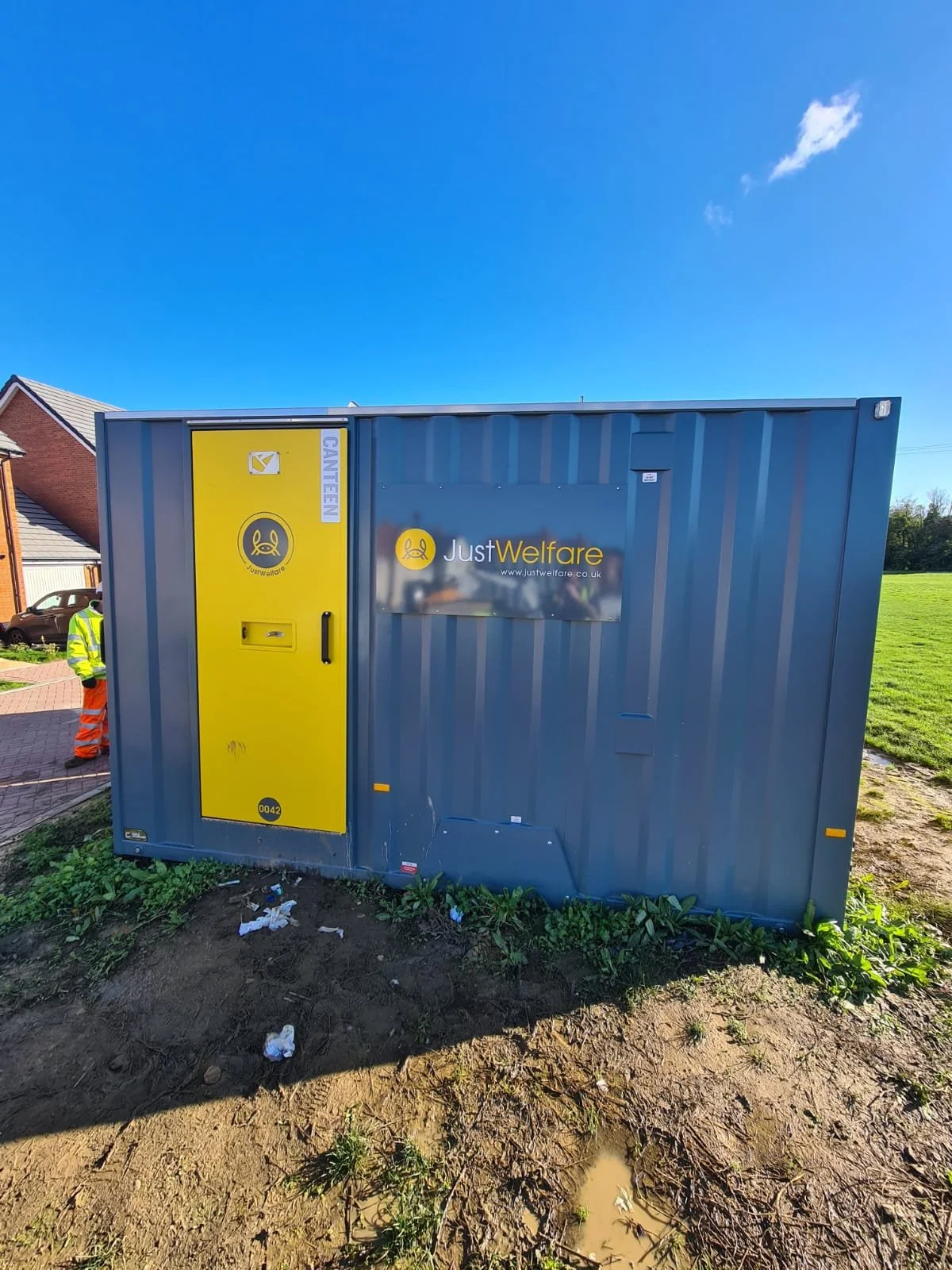 A gray and yellow portable welfare or utility building with a logo and the website 'www.justwelfare.co.uk' on it, situated outdoors on a patch of dirt and grass, with a blue sky in the background.