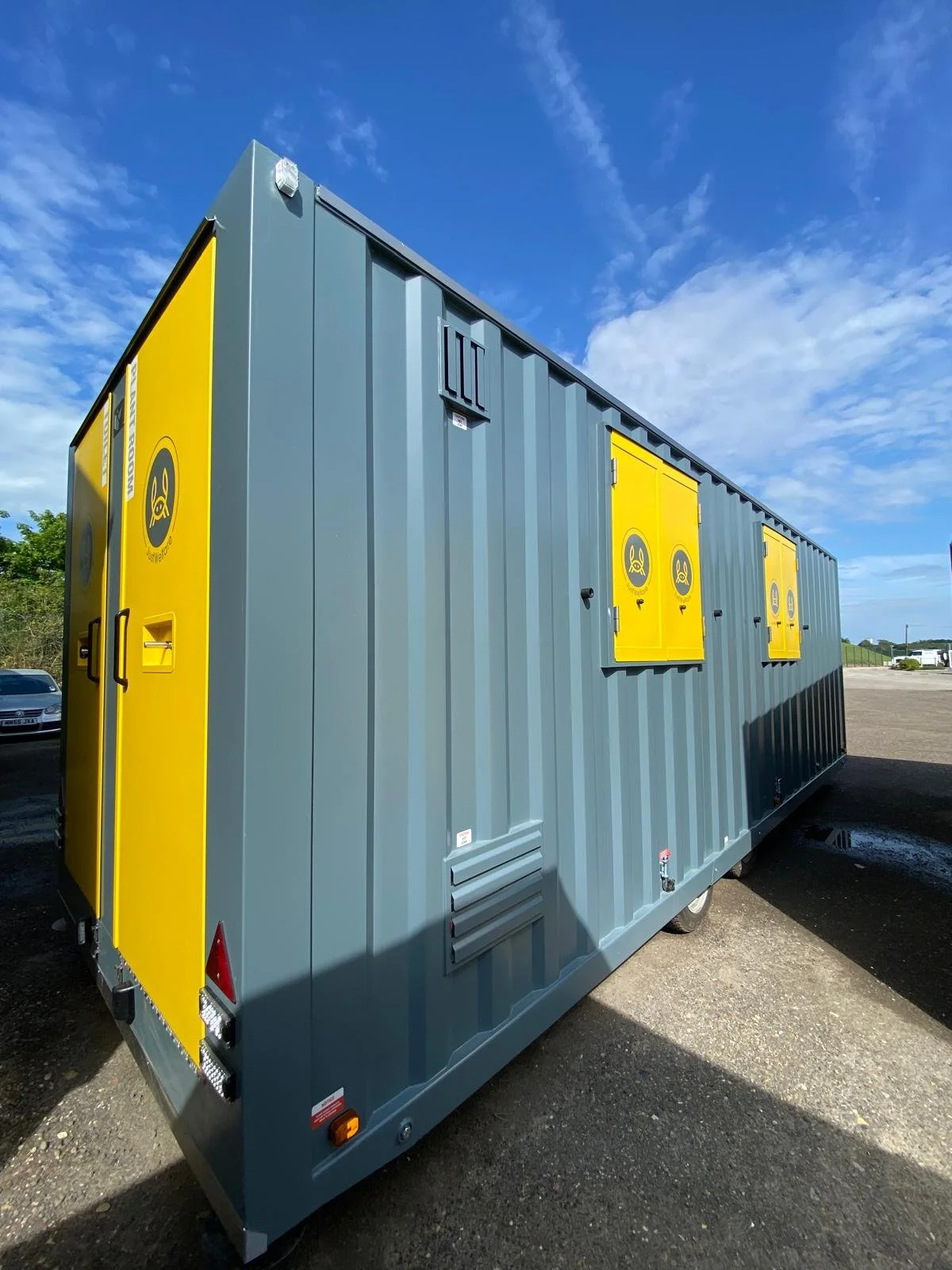 A large gray mobile electrical generator or transformer with yellow warning panels and doors, parked outdoors under a blue sky with some clouds.
