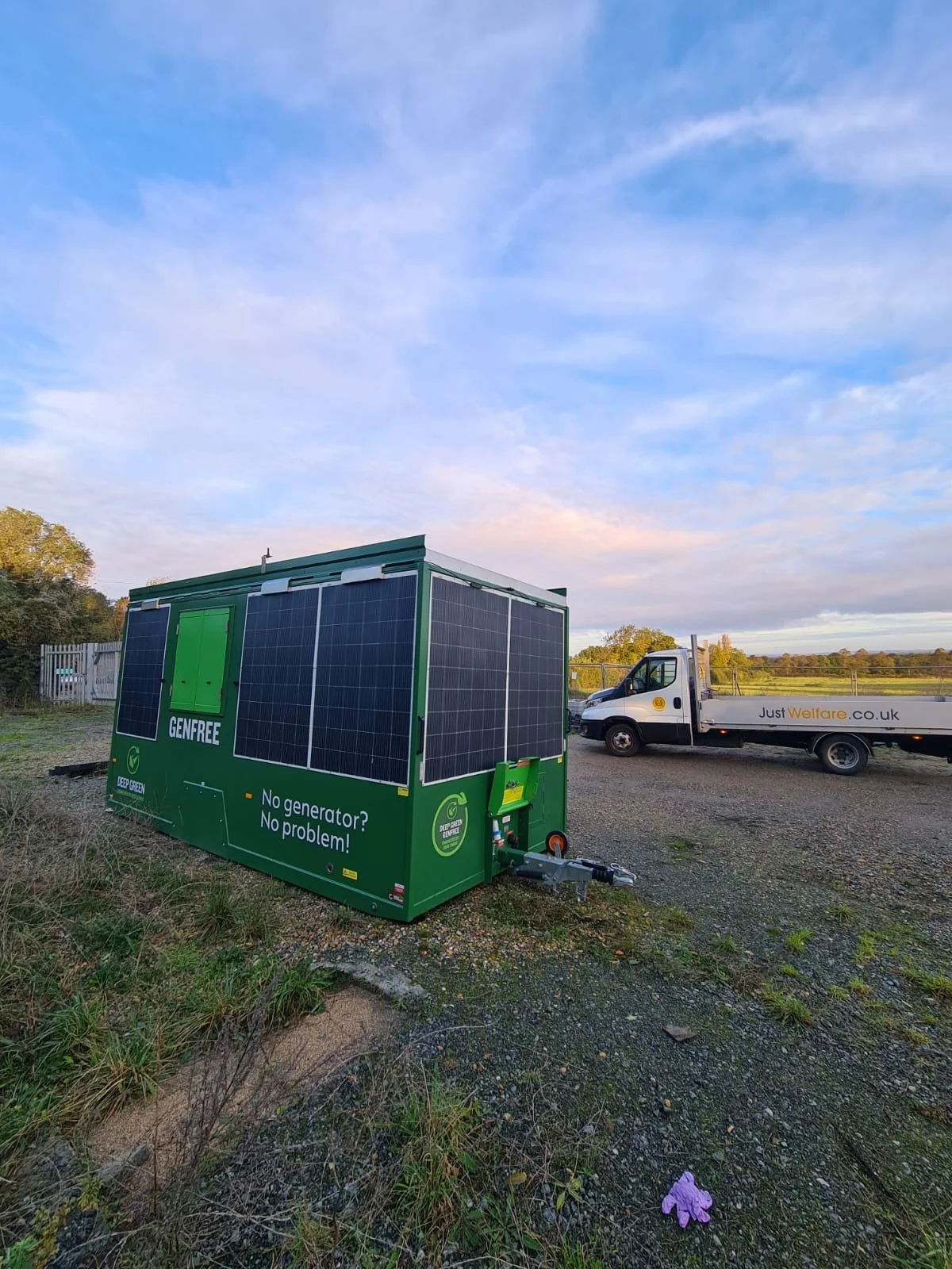 A portable green energy generator with solar panels, labeled 'GENFREE' and 'No generator? No problem!' on a grassy area with a gravel surface, with a pickup truck in the background under a partly cloudy sky.