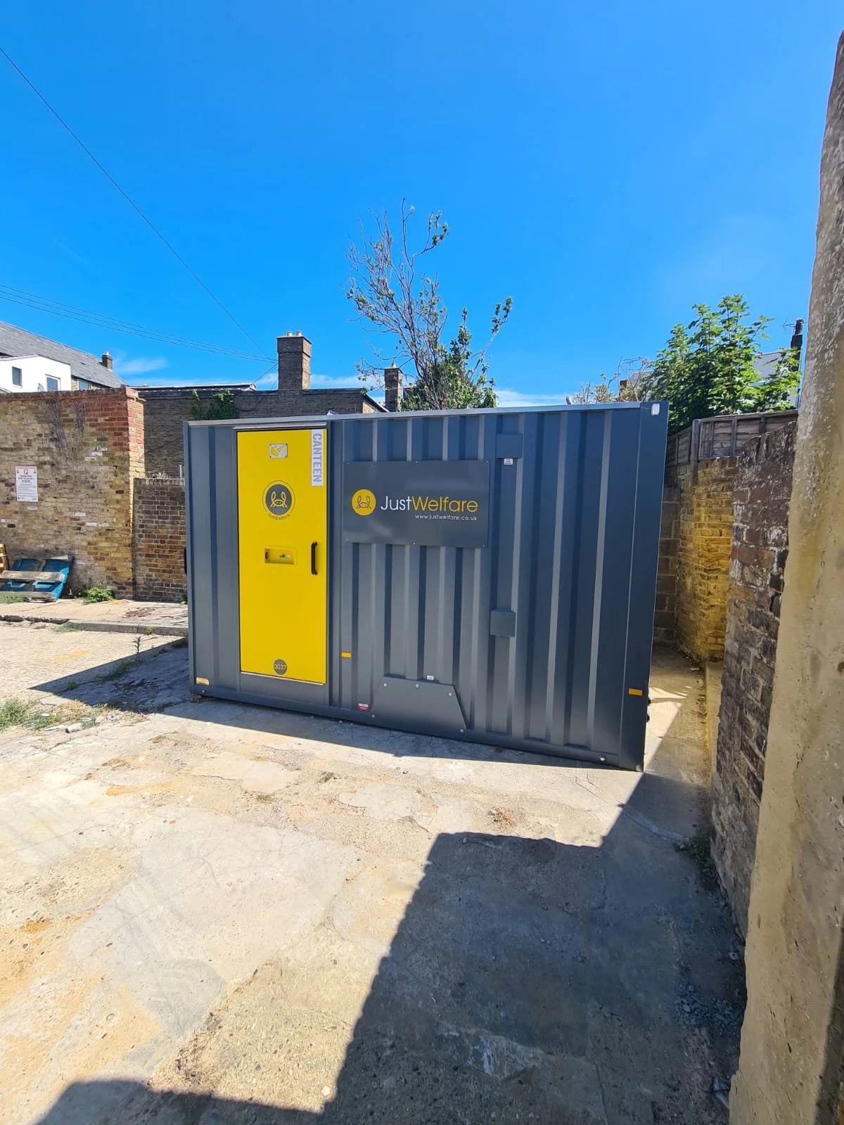 A grey and yellow Just Welfare container with a secure door, placed outdoors on a concrete surface, with an old brick wall and a tree with budding leaves in the background under a clear blue sky.