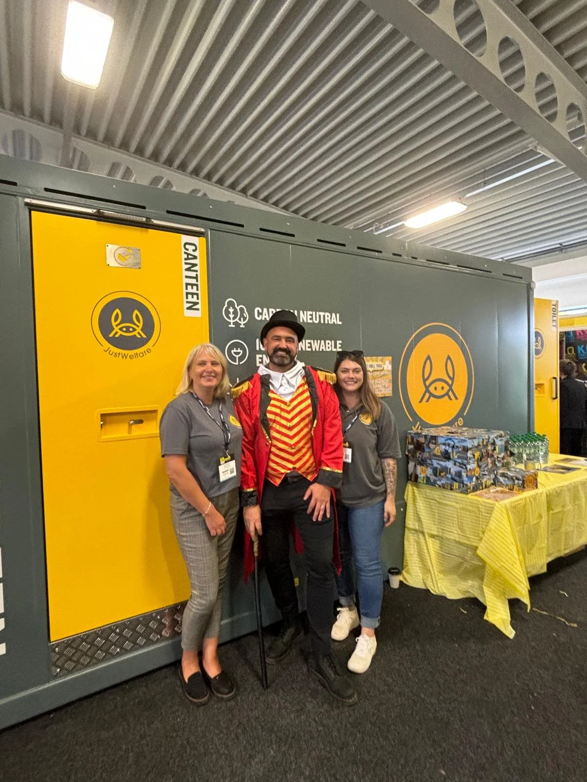 Three people dressed in costumes and casual clothing pose in front of a waste disposal station at an event. The station has a yellow and gray color scheme with signs about waste sorting and environmental themes. One person is dressed as a circus ringmaster with a red and gold jacket, black pants, and a top hat, flanked by two women in gray shirts and checkered pants.