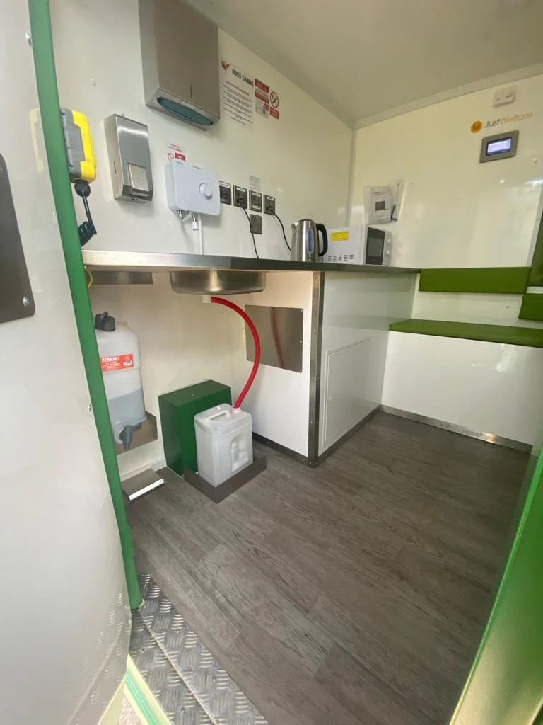 Small kitchen or kitchenette with a stainless steel sink, microwave, electric kettle, and various electrical outlets, with safety signs on the wall.