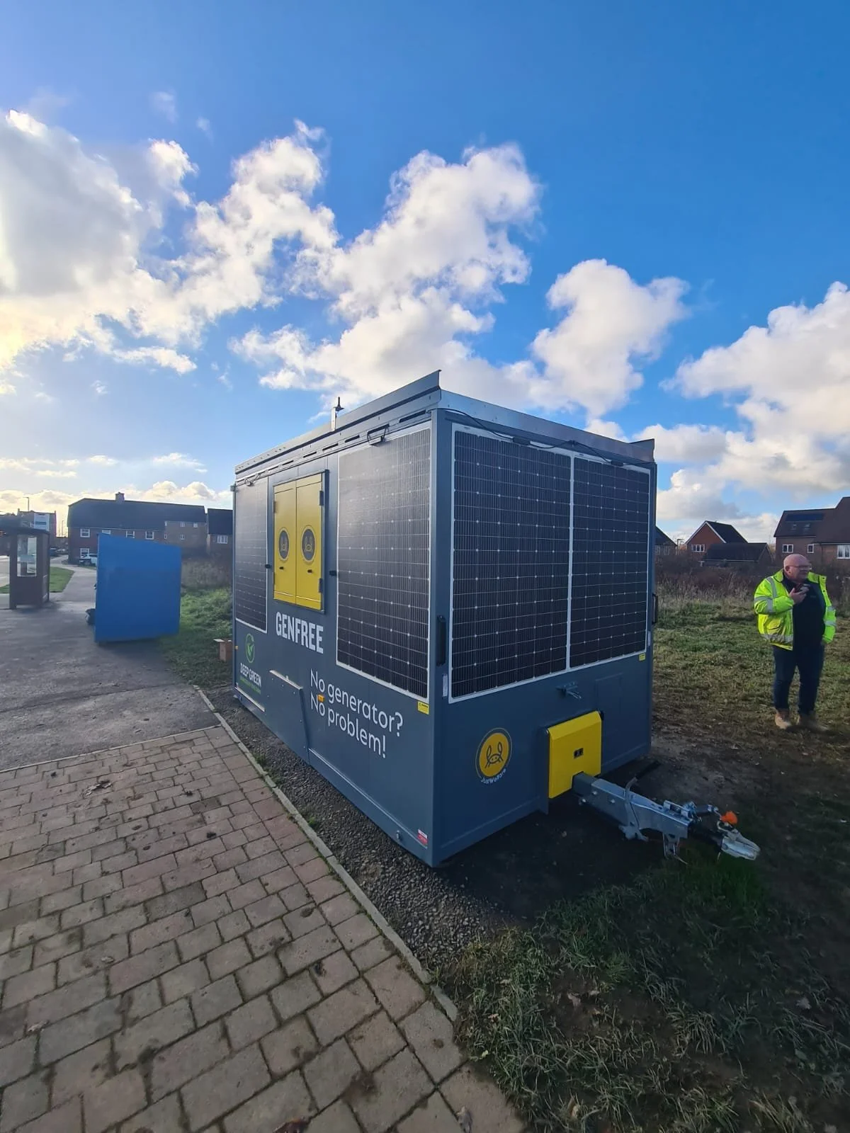 Outdoor energy storage container with solar panels, labeled 'GENFREE' and 'No generator? No problem!', on a paved sidewalk near a grassy area, with a man in a high-visibility jacket nearby and houses in the background under a partly cloudy sky.