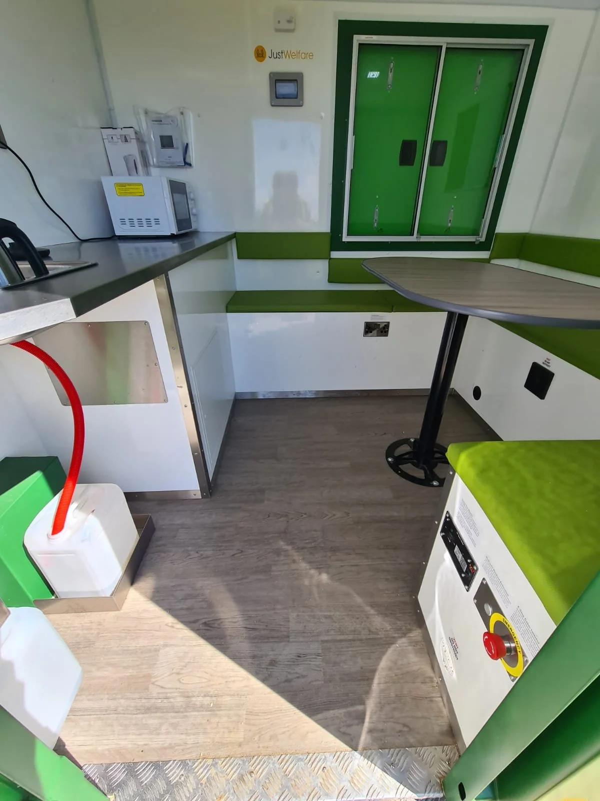 Interior of a small break room or kitchenette with green and white color scheme, featuring a microwave, a wall-mounted telephone, a table, and a green storage cabinet, with wooden flooring.