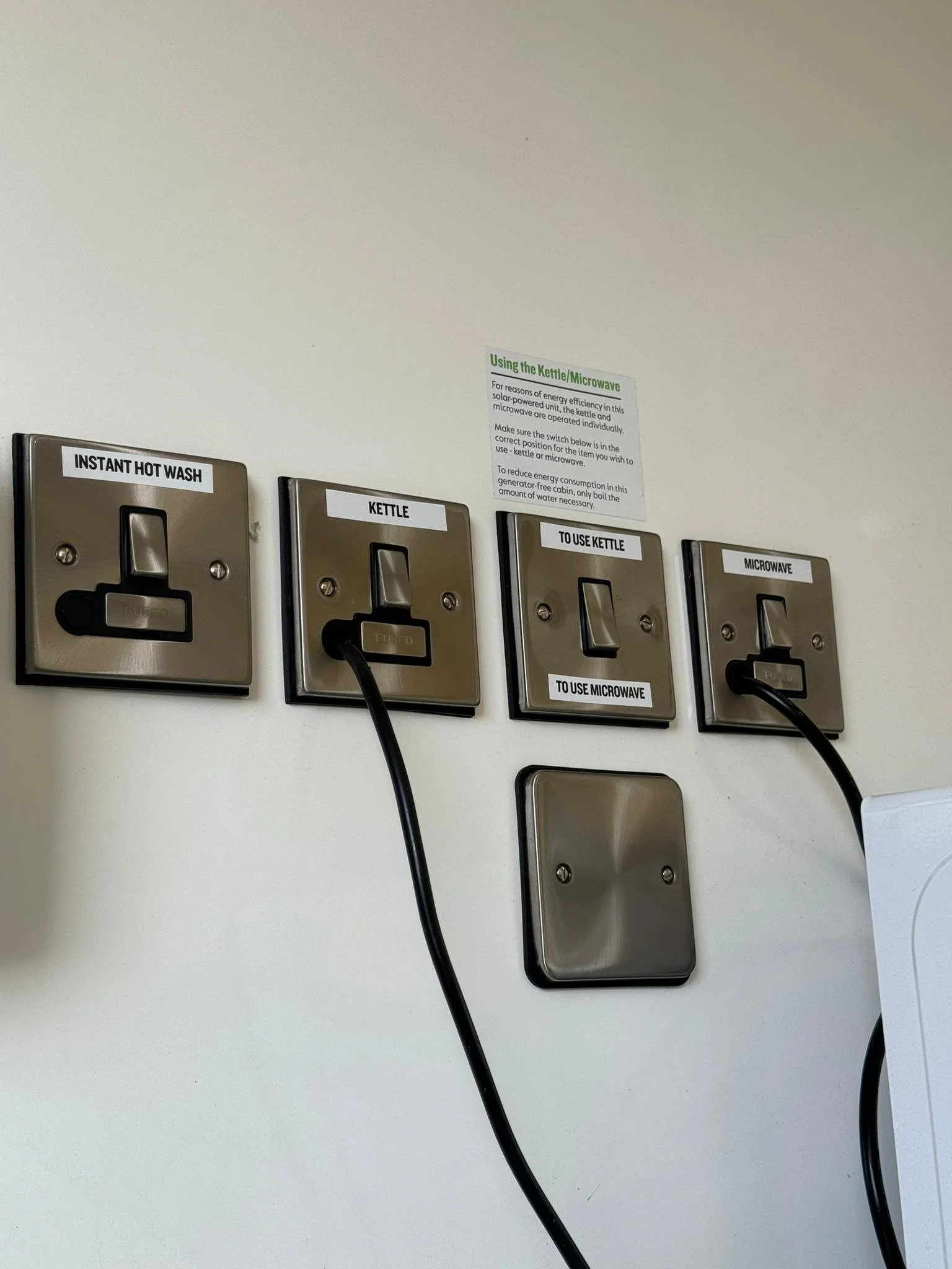 Wall with four electrical switches and outlets labeled for hot water, kettle, microwave, and to use kettle or microwave, with two cords plugged into the hot water and microwave outlets.