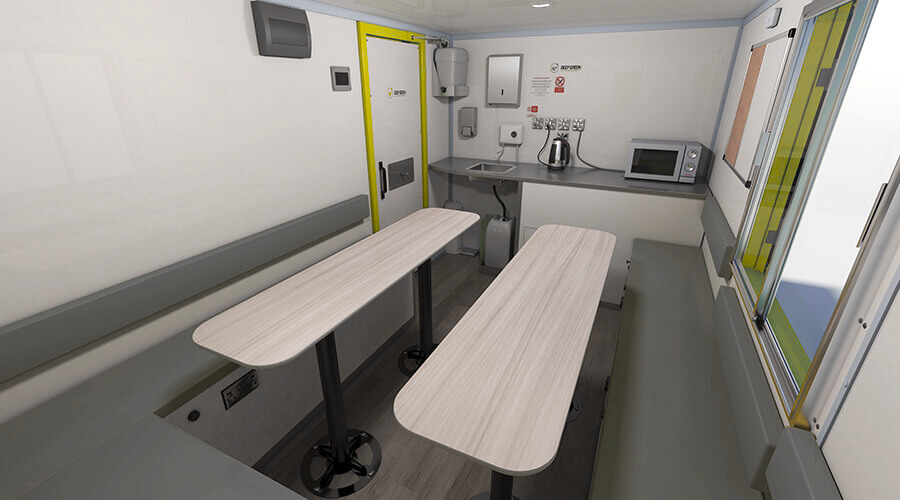 Small, sterile break room with two light-colored tables, a window, a microwave, a kettle, and safety signs.