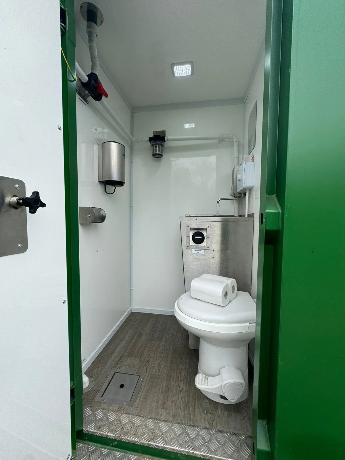 Small, clean, and secure sanitary facility with a white toilet, two rolls of toilet paper on top, and minimal fixtures, including a waste bin, a soap dispenser, and a light on the ceiling.