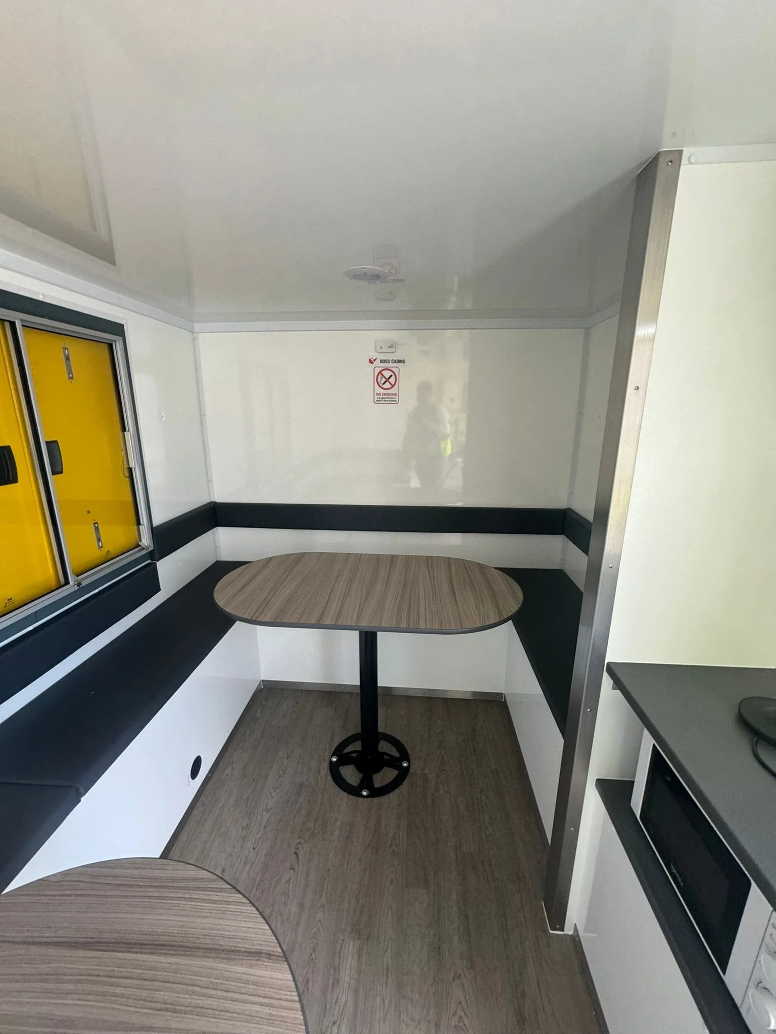 Small dining area inside a mobile or temporary structure with two wooden tables, one partially visible, a microwave oven, and a yellow mailbox or storage box.