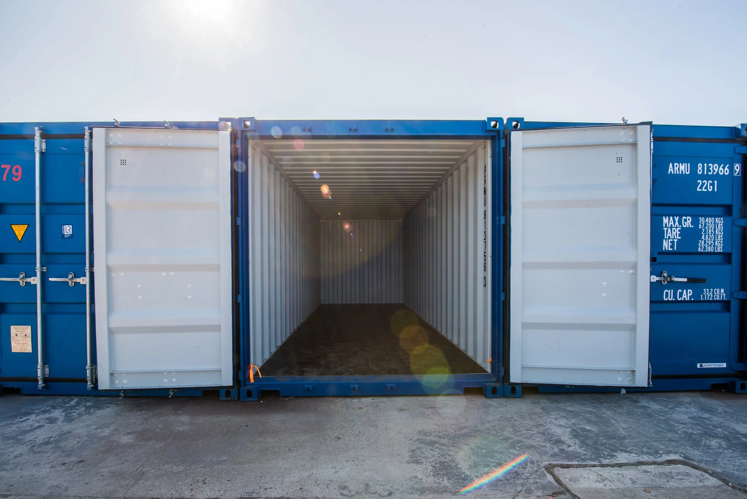 Empty blue shipping container with open doors, sitting on a concrete surface with the bright sun behind it and light flares from the sunlight.