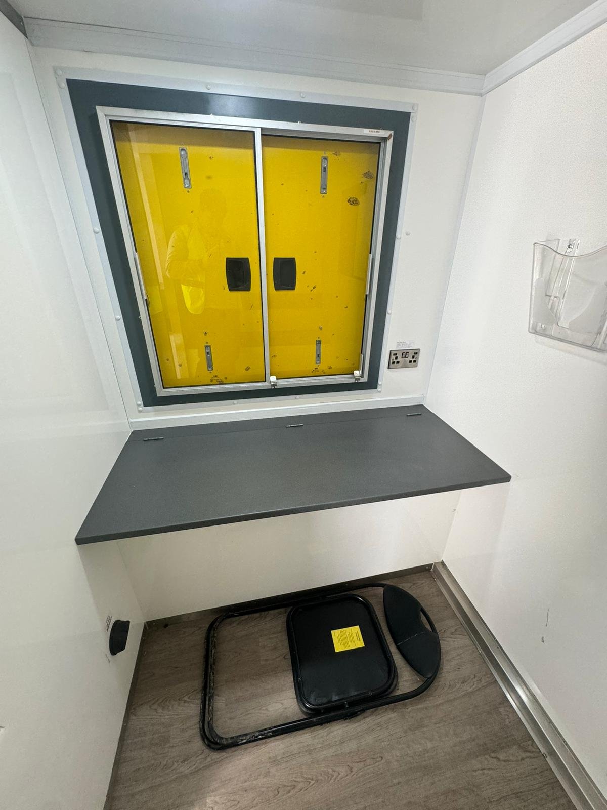 View of an emergency medical supply room with a yellow emergency supply box inside a secure cabinet, a gray countertop, and a black medical emergency kit on the wooden floor.