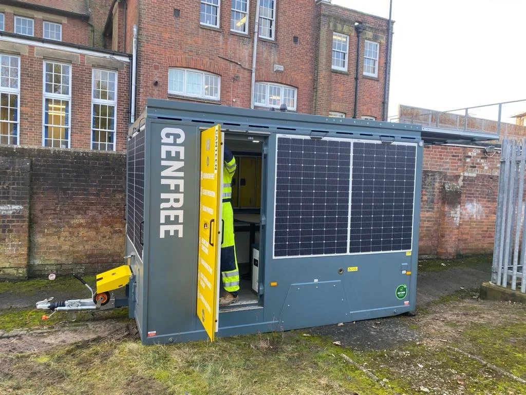 Portable generator covered with solar panels, with a person in a high-visibility jacket entering it, situated outdoors near a brick wall and buildings.