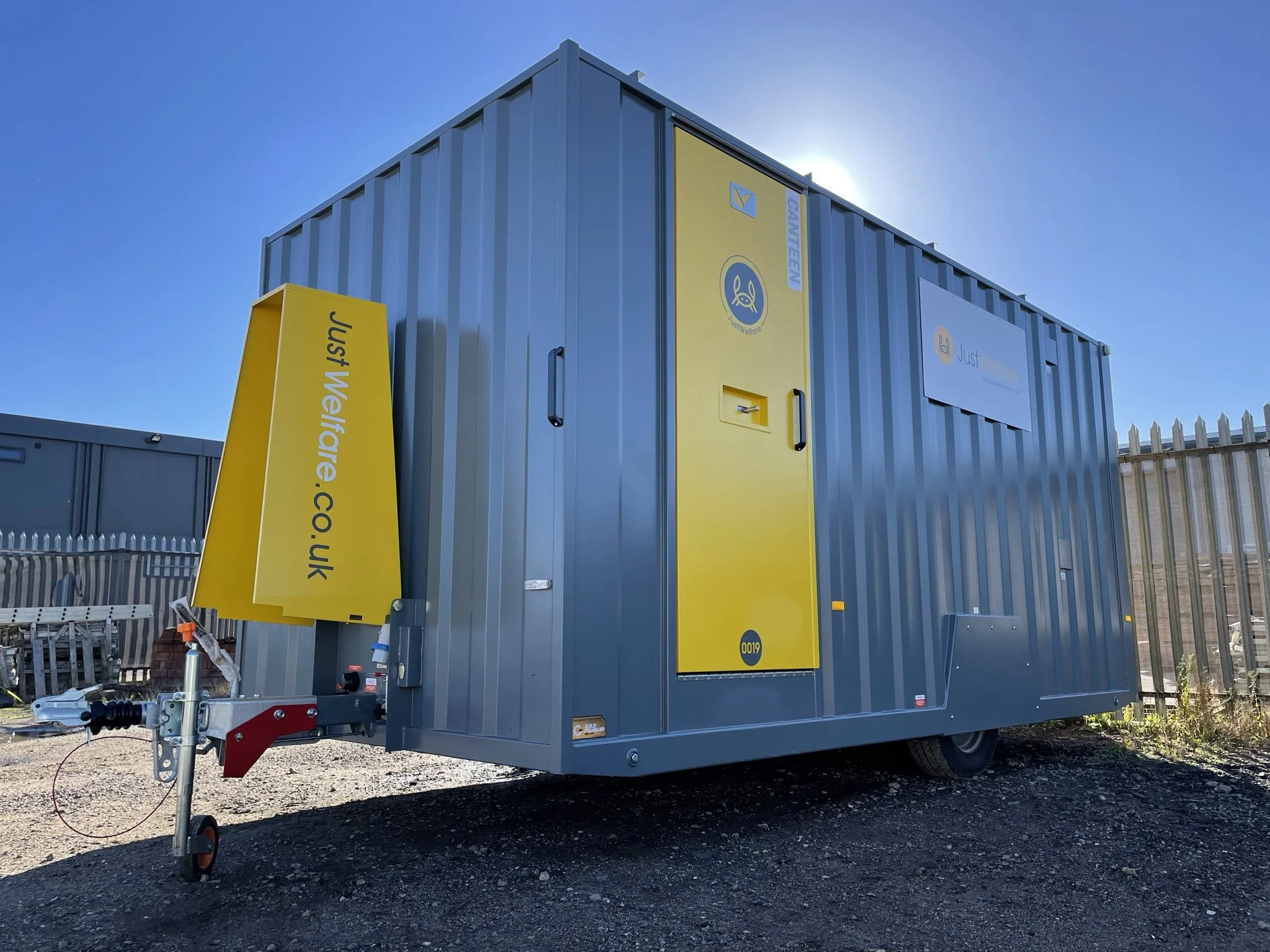 A large grey mobile trailer with a yellow door and yellow sign that reads 'Just Welfare.CO.UK'. It is parked on a gravel lot behind a metal fence.