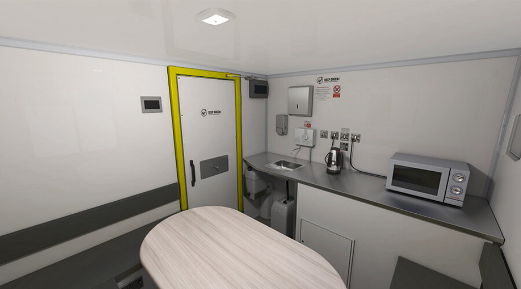 Small medical or scientific room with a wooden table, a microwave on the counter, a kettle, a sink, and various safety and equipment signs on the wall.