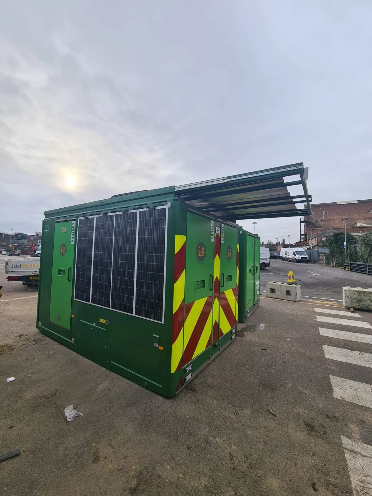 Green utility building with solar panels, construction markings, and a partially open roof, situated outdoors on a construction site with parked vehicles and a cloudy sky.