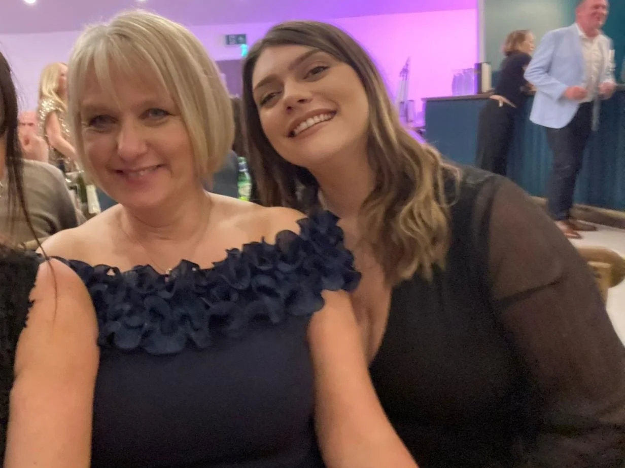 Two women smiling at a social event, with the woman on the left having short blonde hair and wearing a navy dress with floral embellishments, and the woman on the right having long wavy brown hair and wearing a black dress with sheer sleeves.