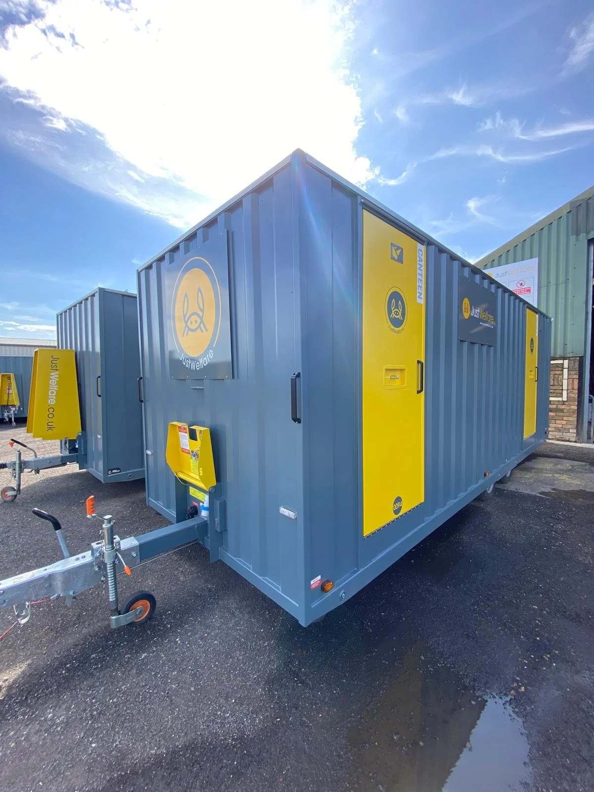 Blue and yellow portable toilets or storage containers with USI Welfare logos, set outdoors on a paved surface, with a bright sky overhead.