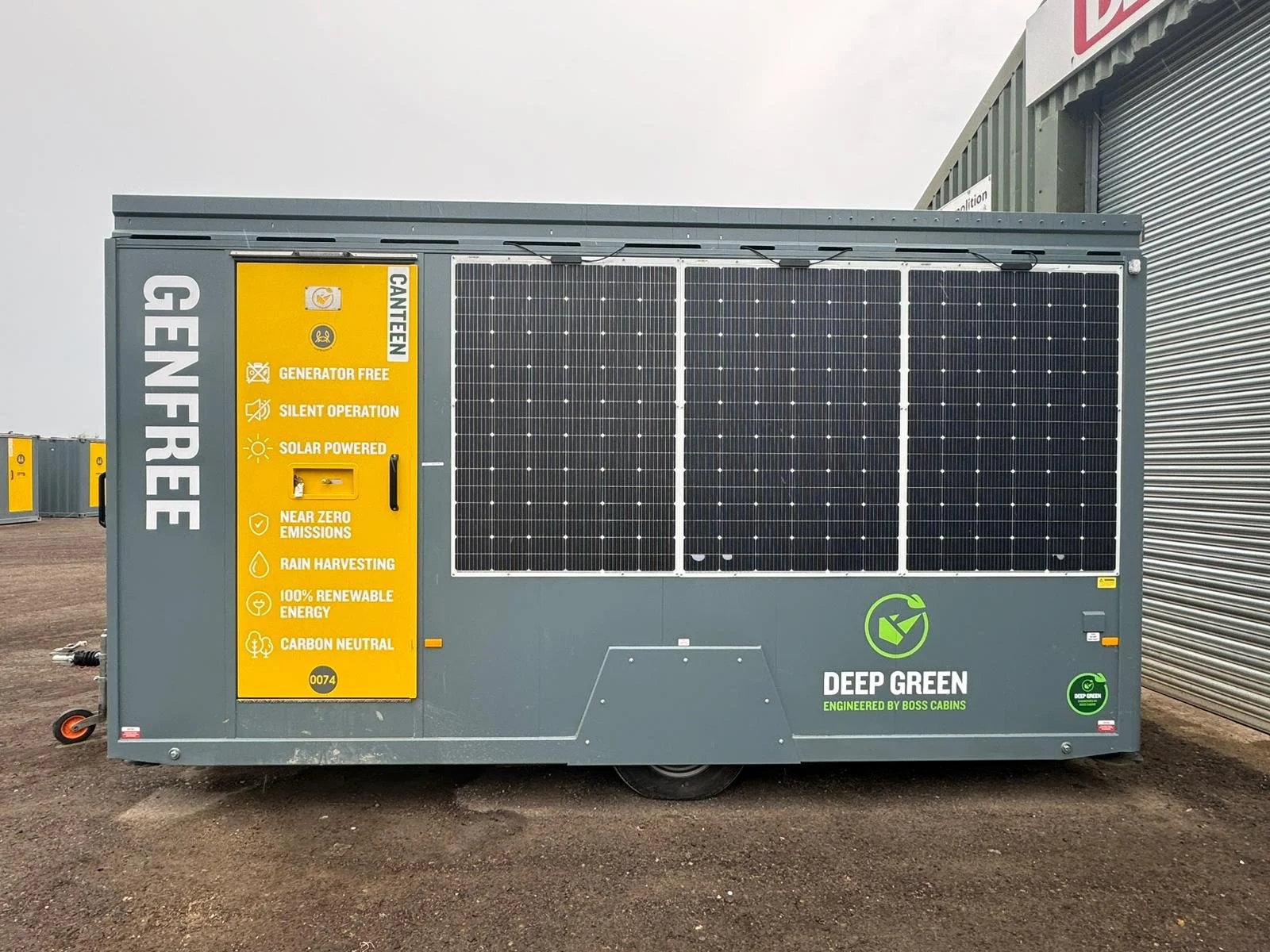 A portable solar power generator with solar panels and yellow informational panel describing its features, located outdoors on a dirt surface with a metal building in the background.