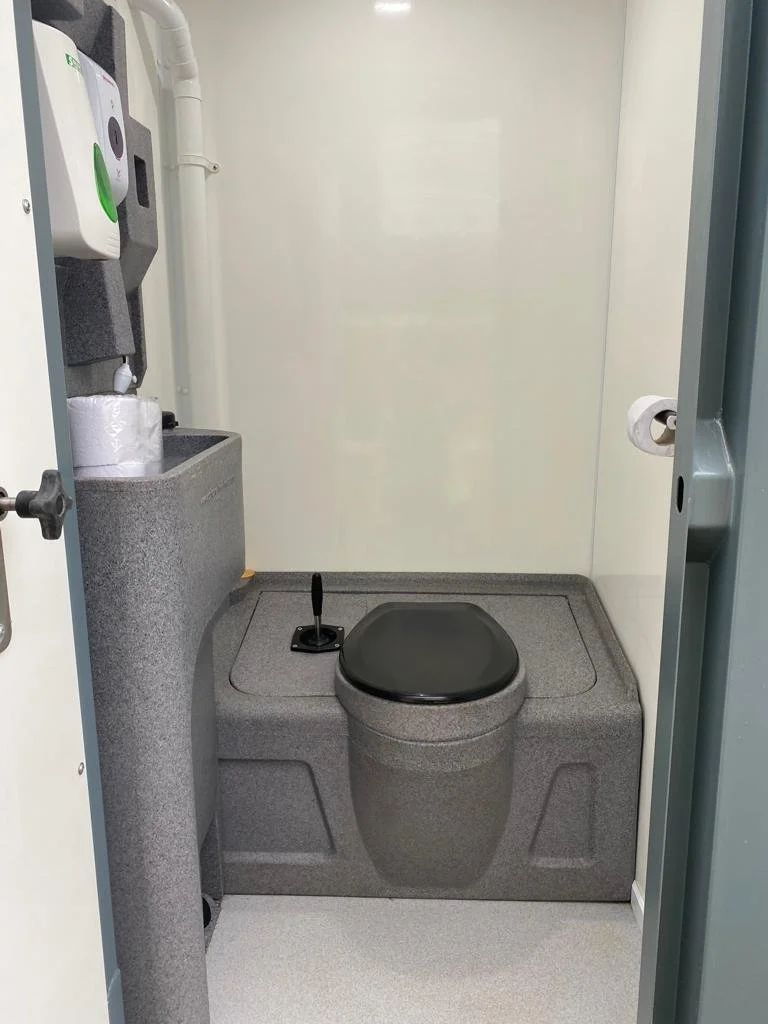 Small, secure bathroom with a gray toilet and black seat, a toilet paper roll, and a sink with a hand sanitizer dispenser.