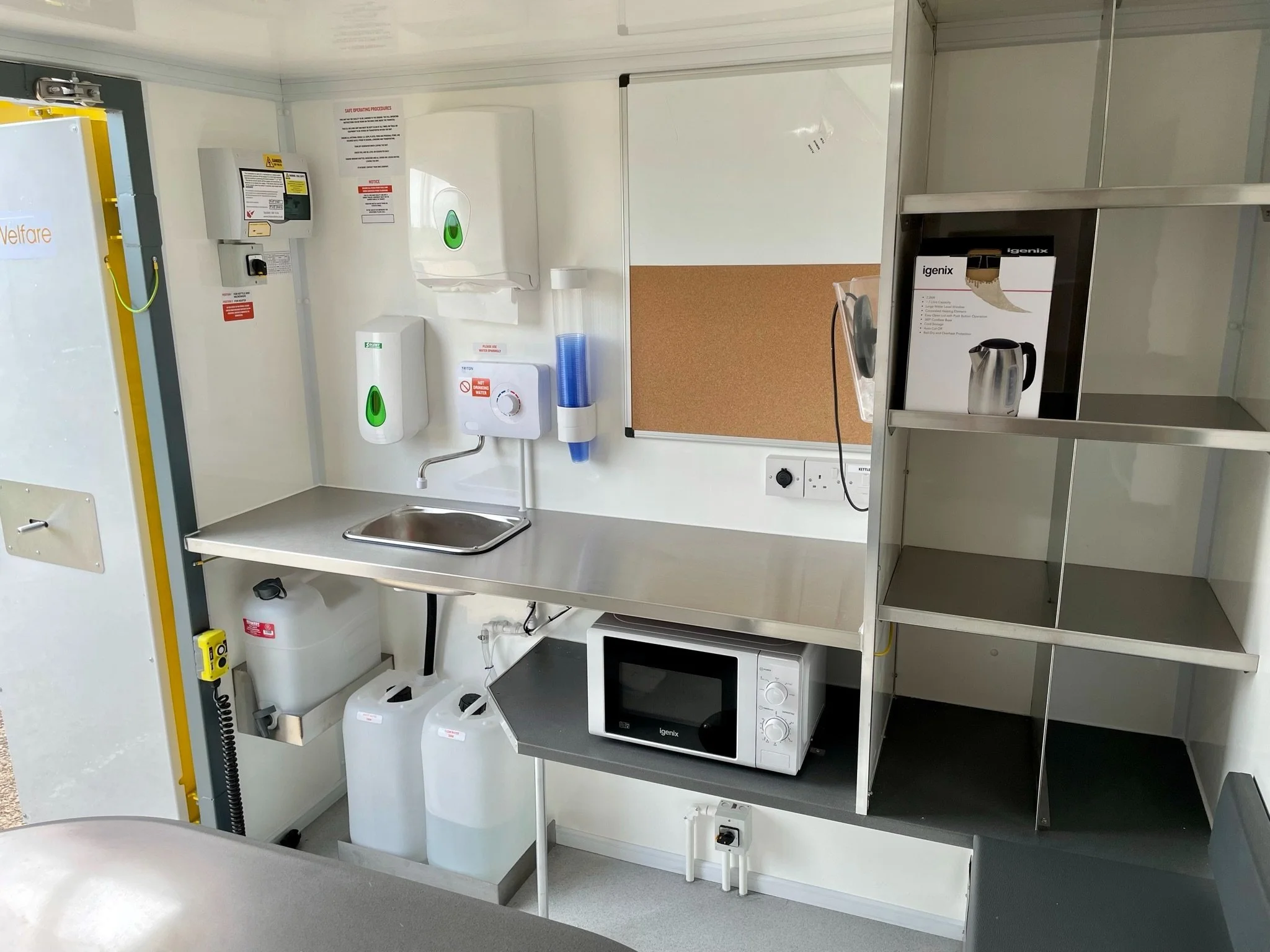 Clean hospital kitchenette with microwave, electric kettle, hand sanitizer, soap dispenser, paper towel dispenser, and storage shelves.