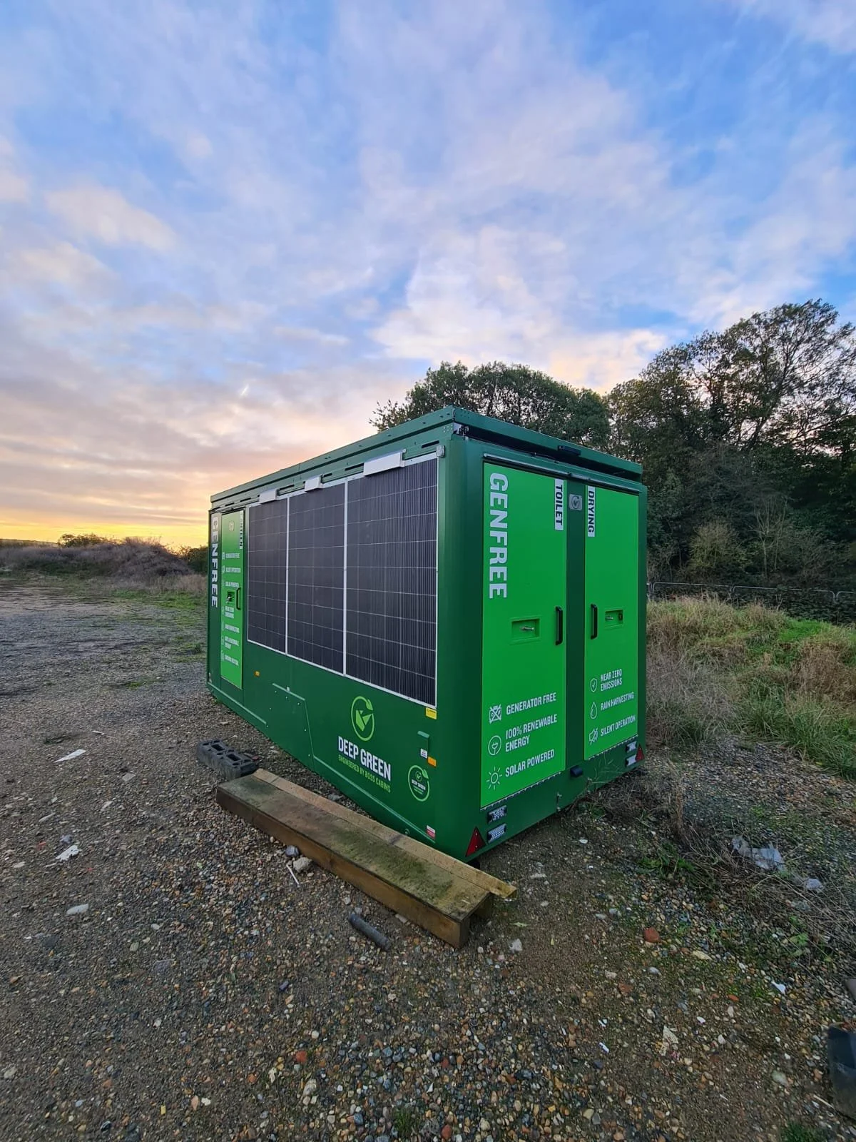 A green portable generator or power storage unit with solar panels on the side, situated outdoors on a gravel surface during sunset, surrounded by trees.
