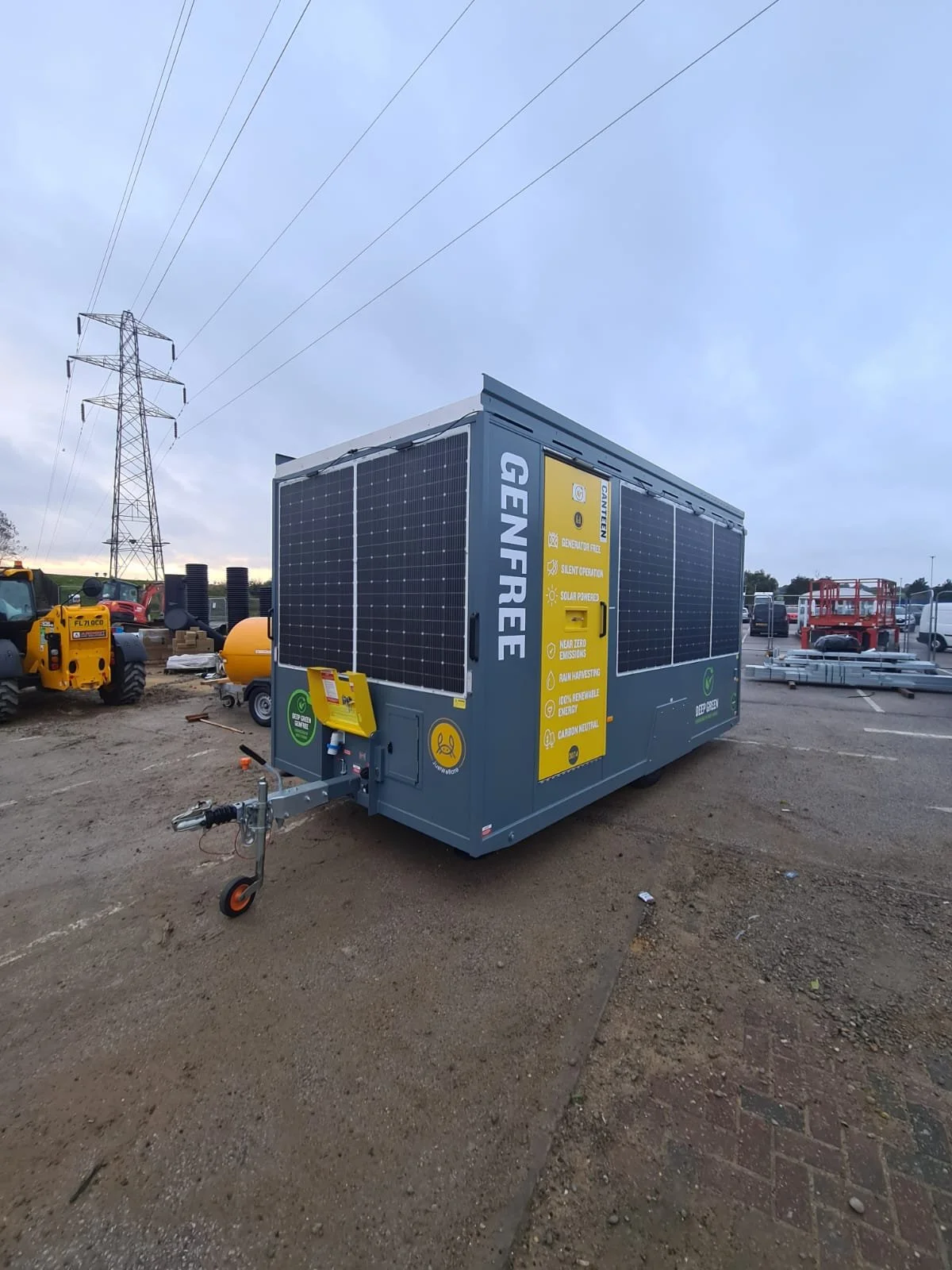 A mobile generator with solar panels installed on its sides, parked on a dirt lot with construction equipment and power lines in the background.