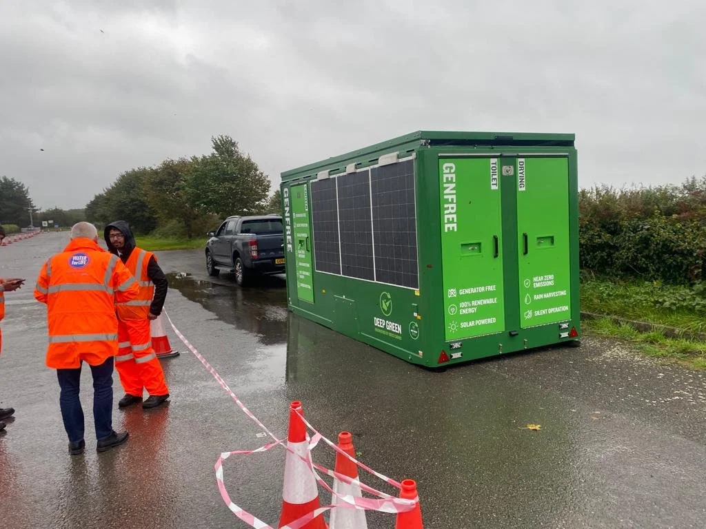 A green portable generator with solar panels on top, labeled 'Green-Free' and 'Deep Green,' is parked on a wet road next to a black truck. Two men in orange safety vests are talking near the generator, and orange traffic cones and tape cordon off the