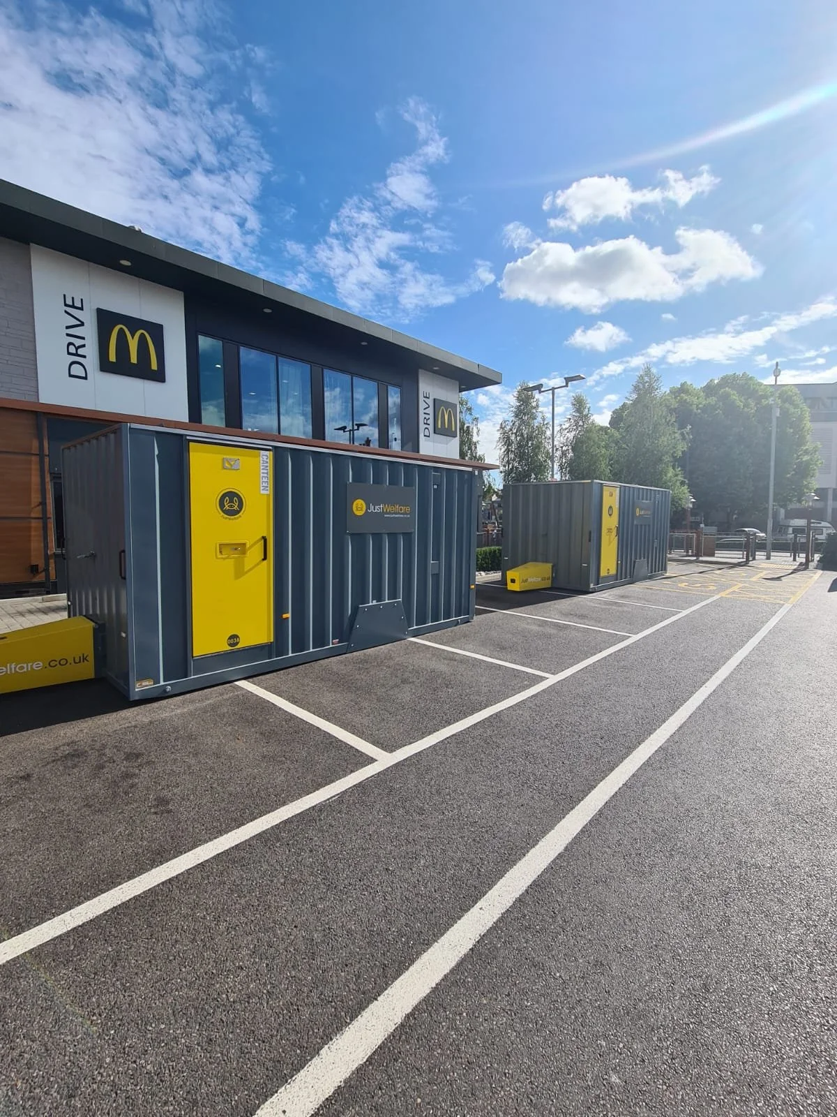 A parking lot outside a building with a McDonald's drive-thru sign, two gray steel containers with yellow logo and markings, and a nearby yellow and gray parcel delivery locker.