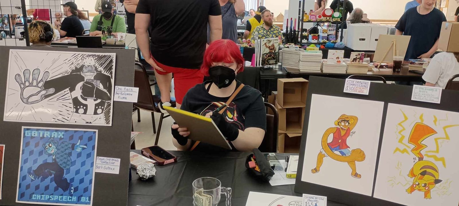 A woman sitting at a table at an art convention, wearing a black mask and gloves, holding a sketchpad, surrounded by various artwork displays, with other people browsing and selling art in the background.