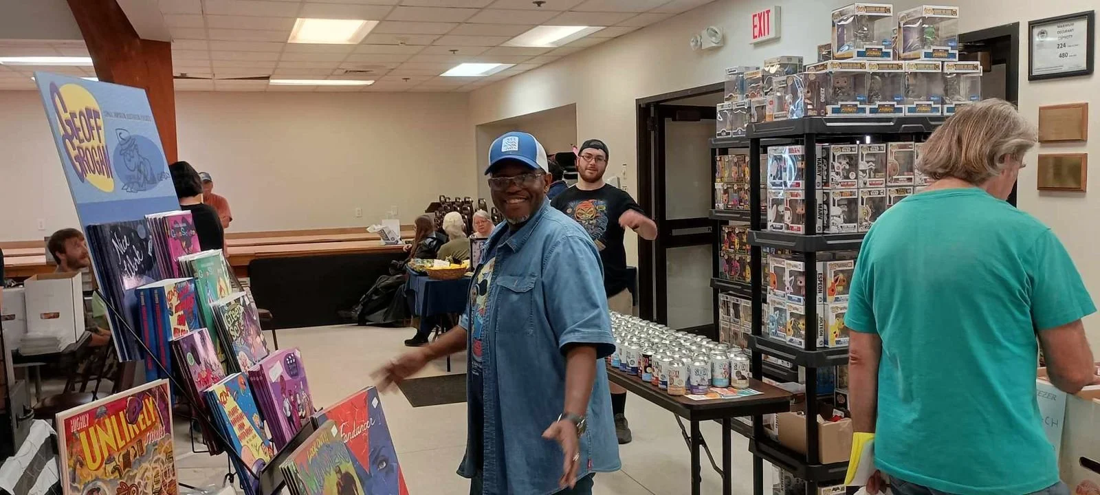A man in a denim shirt and blue cap smiling at a comic book fair, with various shelves of comic books and merchandise around him. Other people are browsing in the background.