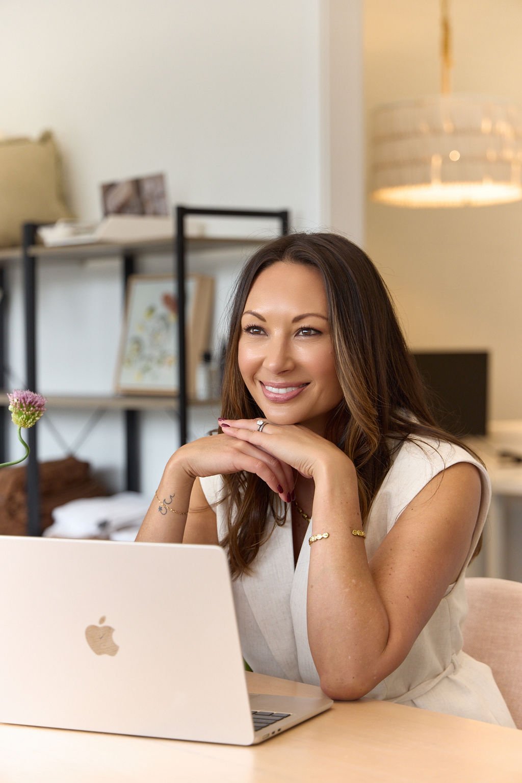 A smiling woman with long brown hair sitting at a desk with an open MacBook, resting her chin on her hands, in a modern home or office space.