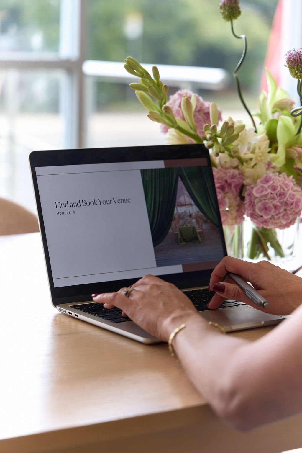 A person using a laptop on a wooden table, with a bouquet of pink and white flowers in a vase nearby, inside a room with a window in the background.